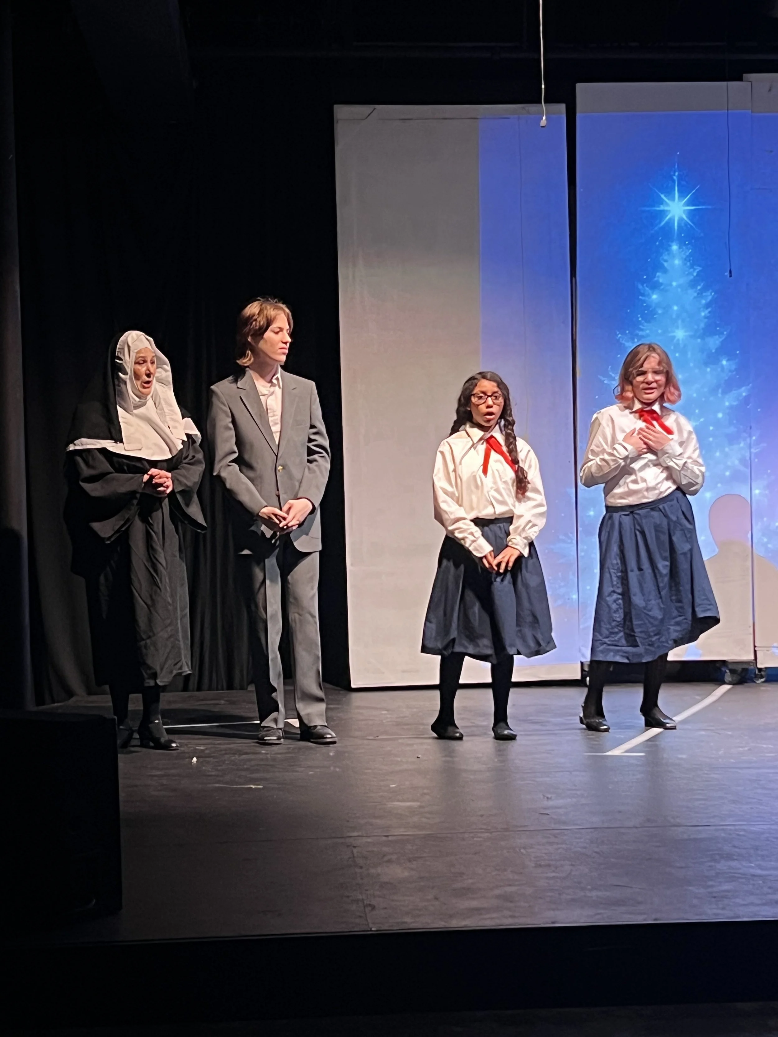 Four actors on stage during a theatrical performance, one dressed as a nun, one in a gray suit, and two in school uniforms with skirts and red neckties, with a blue Christmas-themed background.