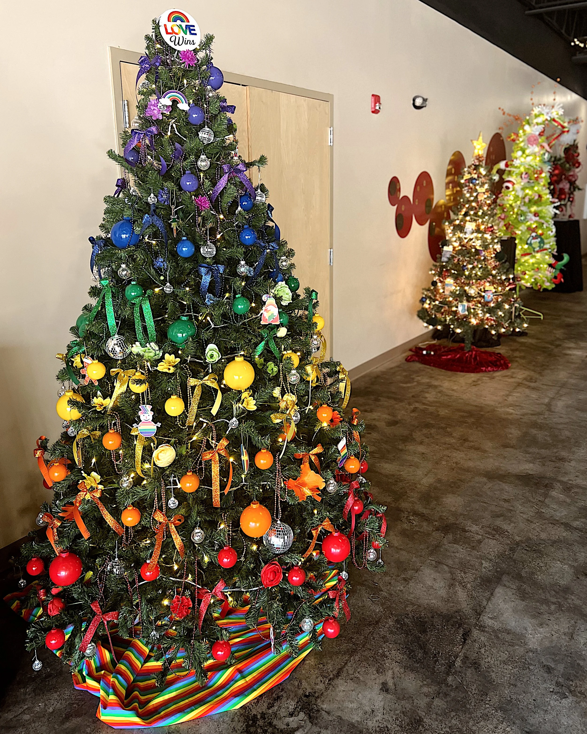 Colorful Christmas trees decorated with ornaments and ribbons, with a rainbow-striped tree skirt at the base, in an indoor setting.