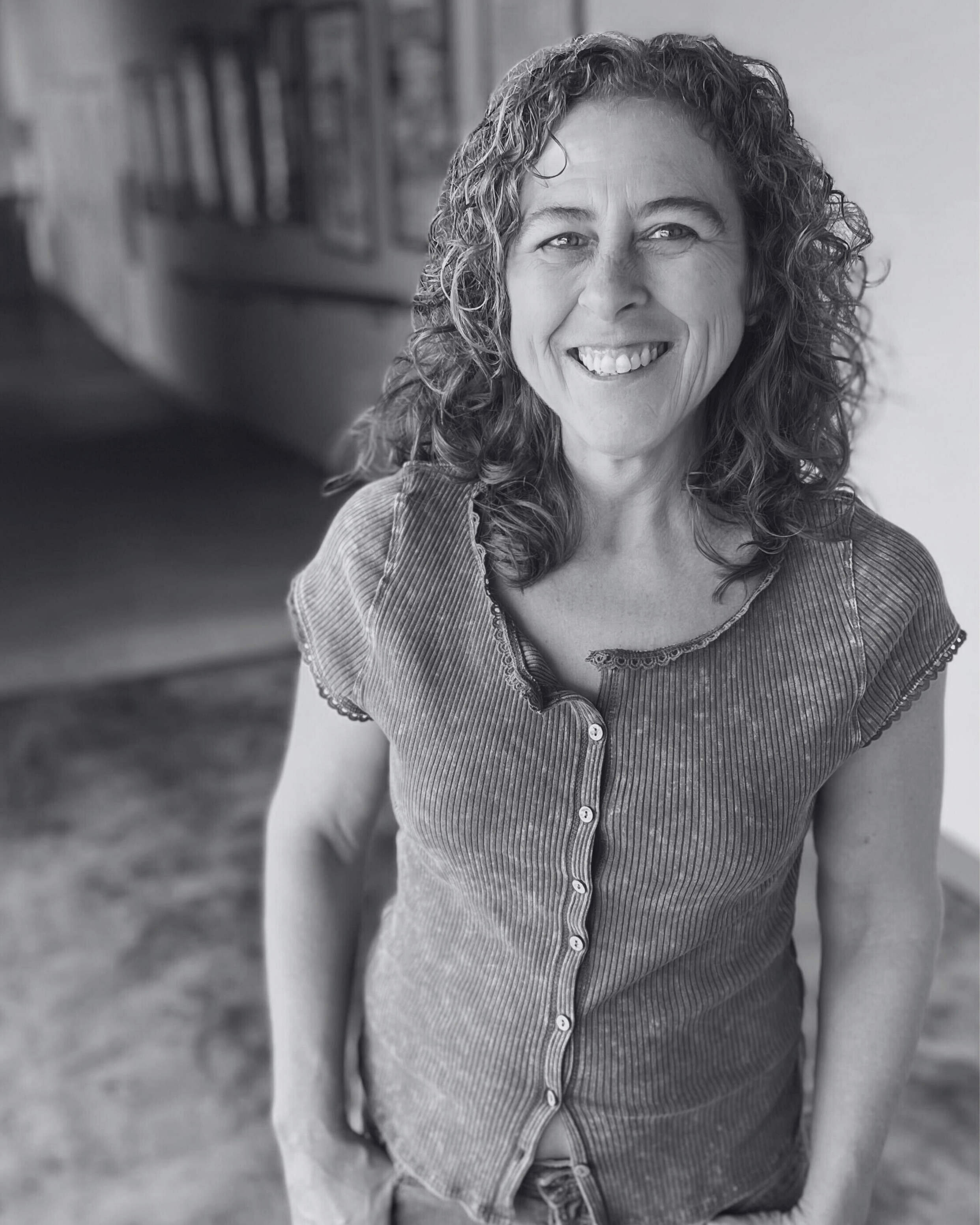 A woman with curly brown hair and a big smile, wearing a purple button-up top, standing indoors with a blurred background of art and books.