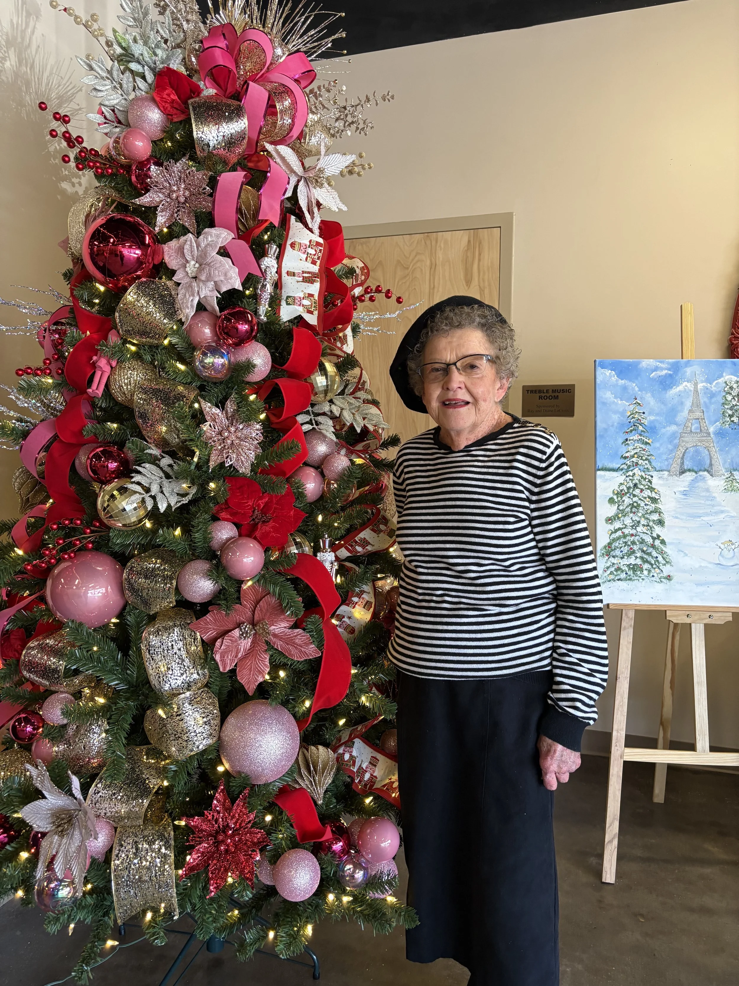 An elderly woman with curly hair and glasses standing next to a decorated Christmas tree with pink, gold, red, and silver ornaments and ribbons, in an indoor setting with a painted winter scene on an easel nearby.