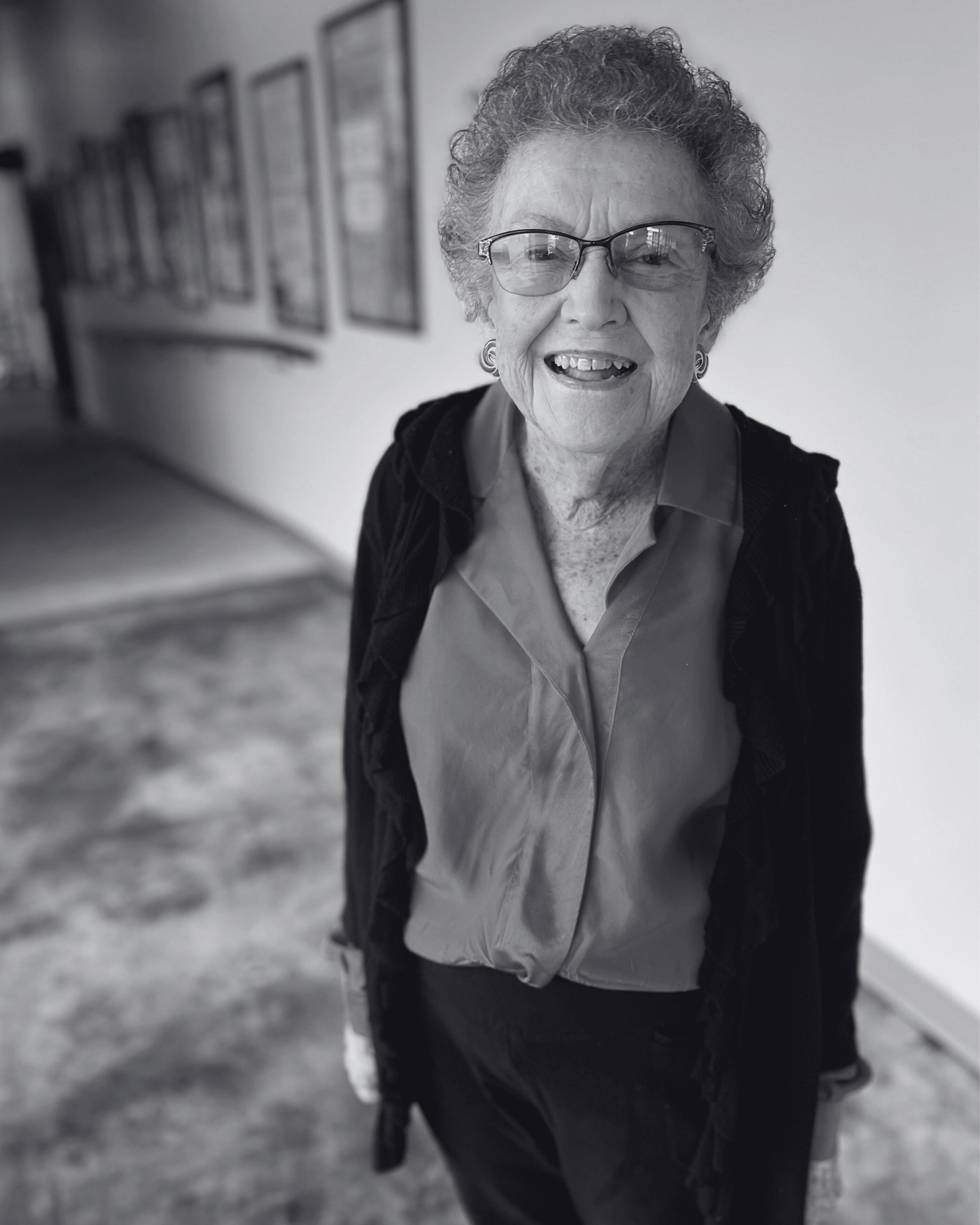 An elderly woman with curly gray hair and glasses, smiling, wearing a red blouse and black jacket, standing indoors with framed pictures on the wall behind her.