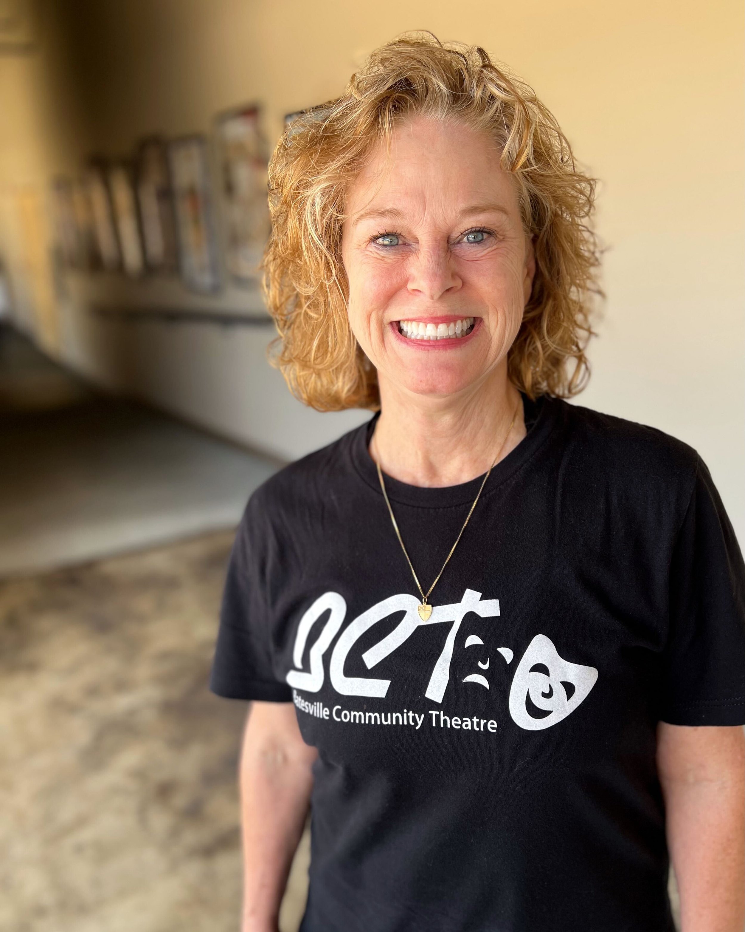 A cheerful woman with curly red hair smiling, wearing a black t-shirt with 'GCT' and 'Gatesville Community Theatre' printed on it, and a gold necklace with a small heart pendant, standing in a hallway with pictures on the wall.