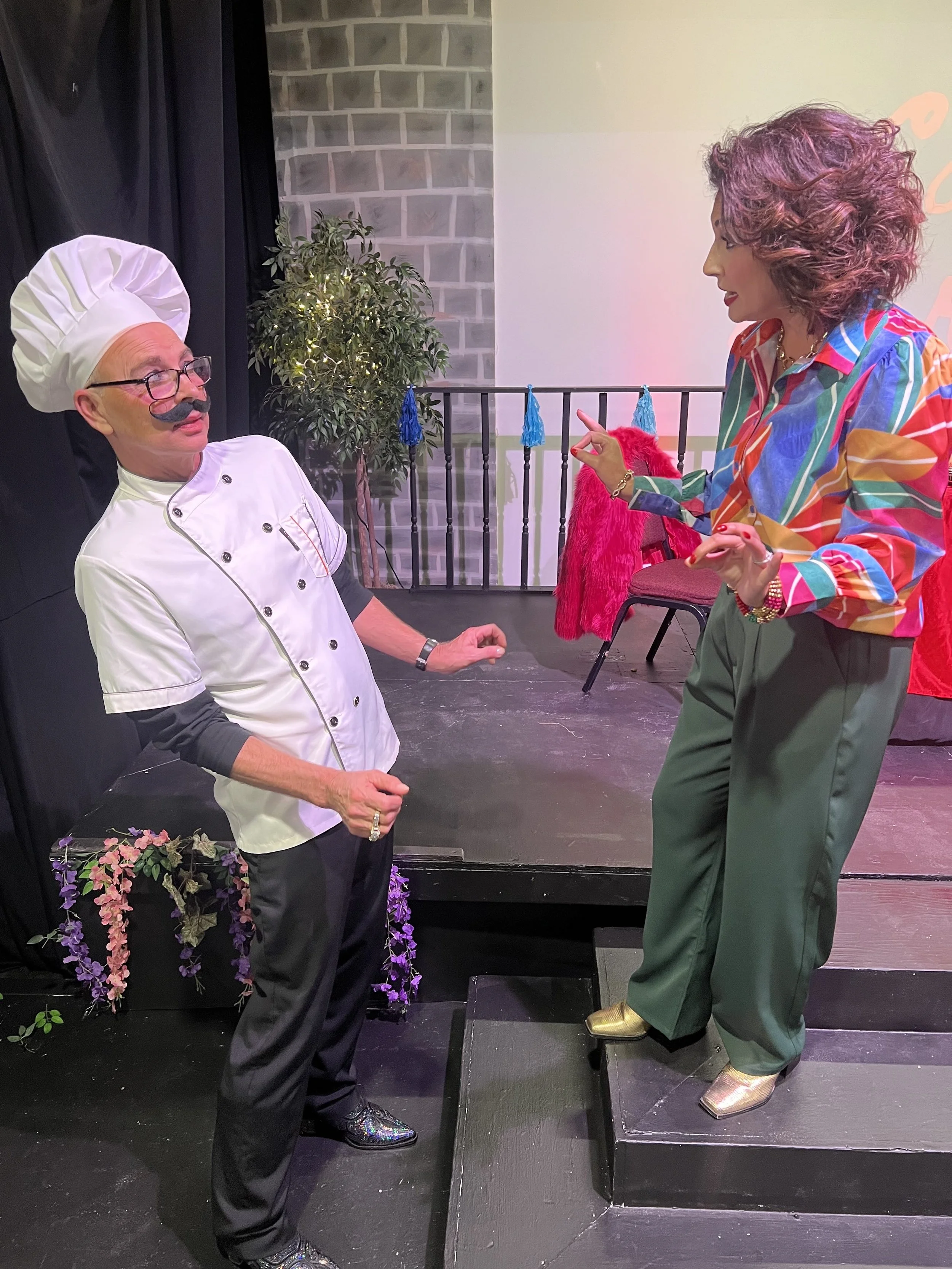A man dressed as a chef with a chef hat, glasses, and a fake mustache is talking to a woman on a small stage. The woman has curly hair, wears a colorful striped shirt, green pants, and gold shoes. The background includes a brick wall, a potted plant, and stage props like a red coat and blue tassels.