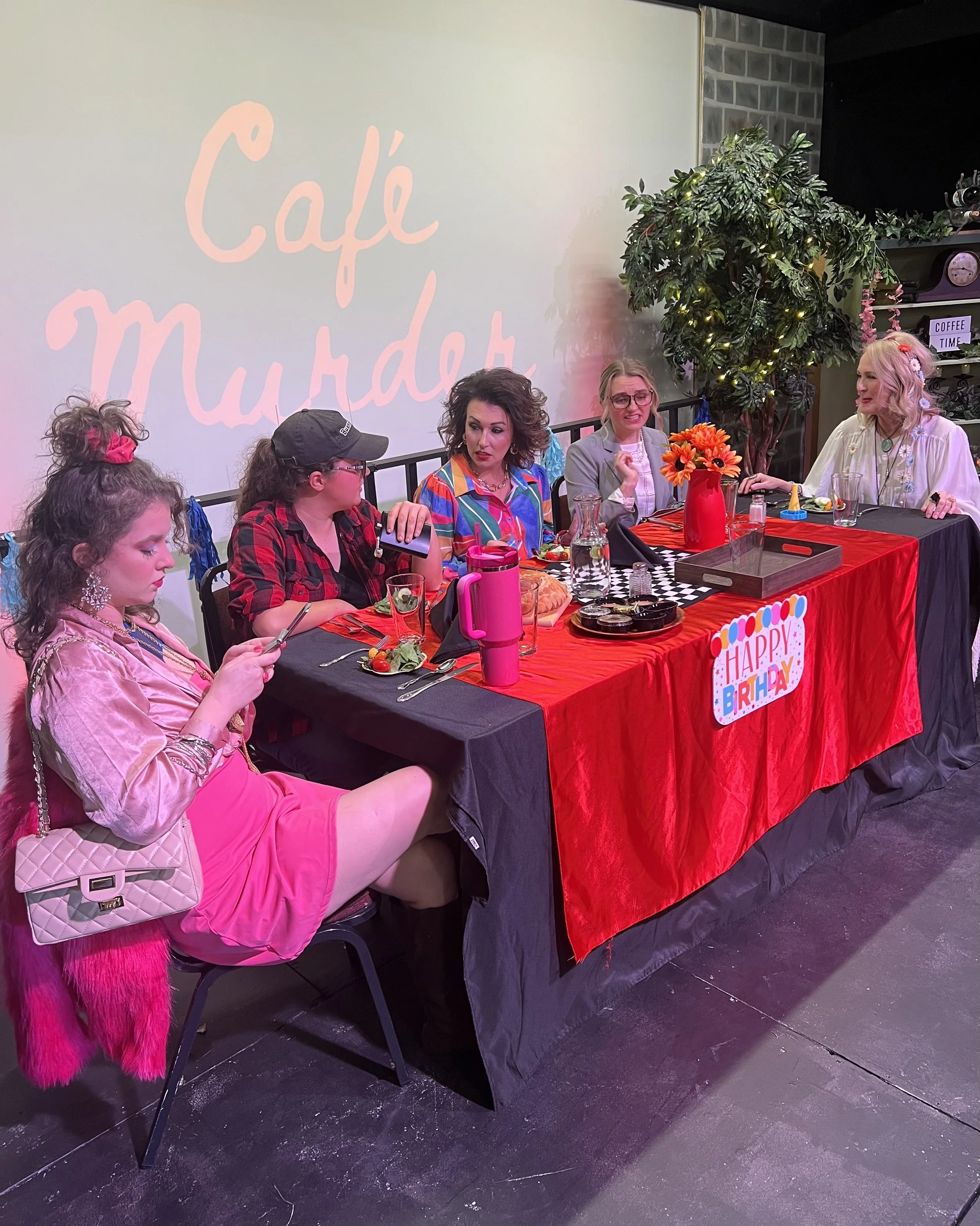 A birthday celebration scene with six women sitting at a table decorated with a 'Happy Birthday' sign, flowers, and various food and drinks, in a room with a large wall projection reading 'Café Murder.'