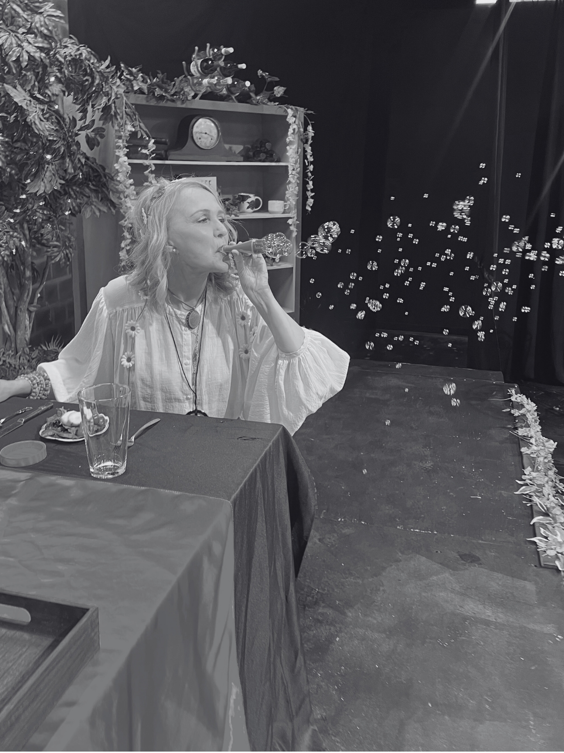 A woman sitting at a dining table, blowing bubbles through a bubble wand, with a decorated background and a table set with a glass and some food.