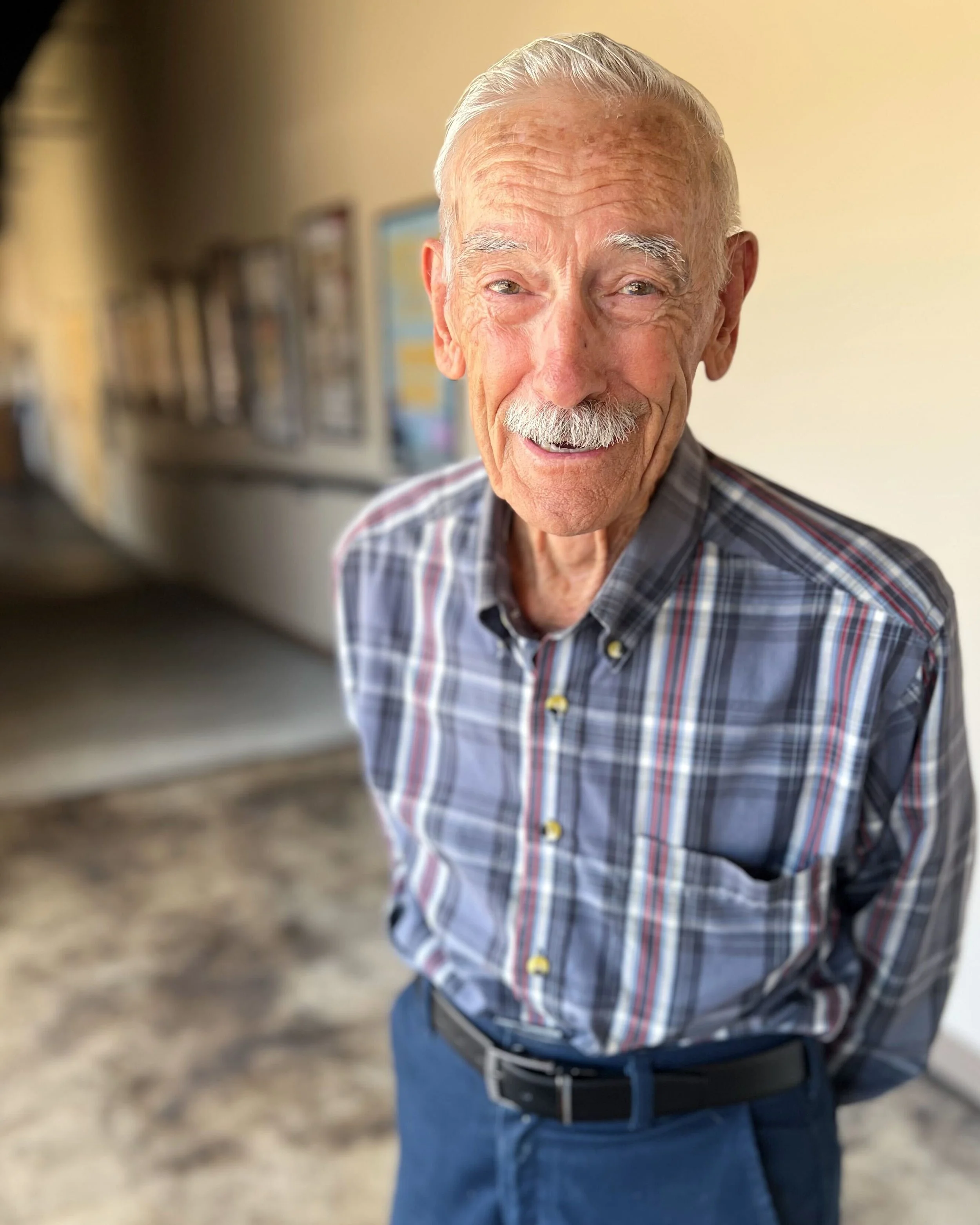 An elderly man with white hair, a mustache, and a friendly smile, wearing a plaid shirt and blue jeans, standing indoors in front of a beige wall and blurred background.