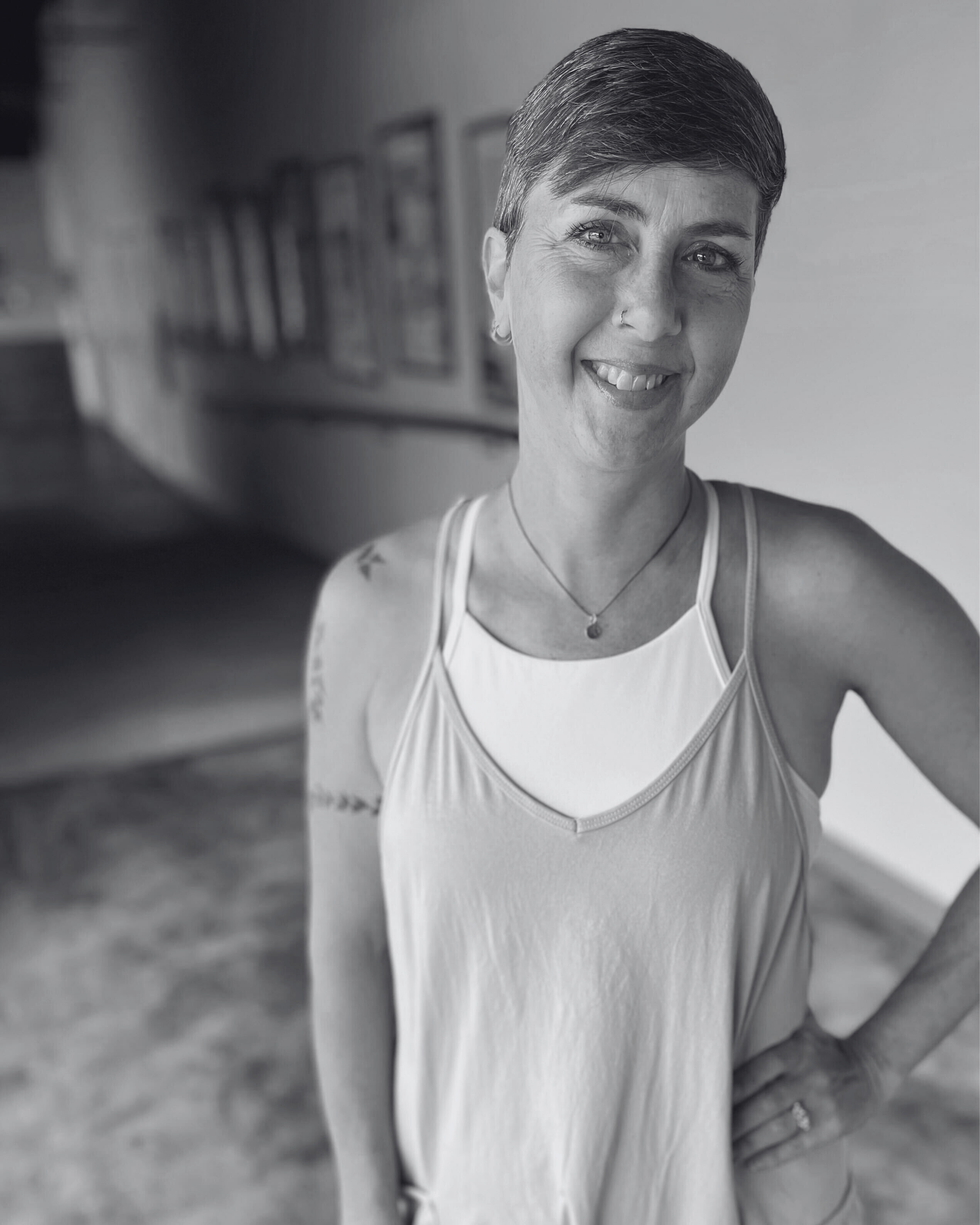 A woman with short hair, blue eyes, and a nose ring taking a selfie. She is wearing a white tank top layered over a white sports bra, a necklace, and has tattoos on her left arm. She is smiling and standing in an indoor setting with blurred artwork on the wall behind her.
