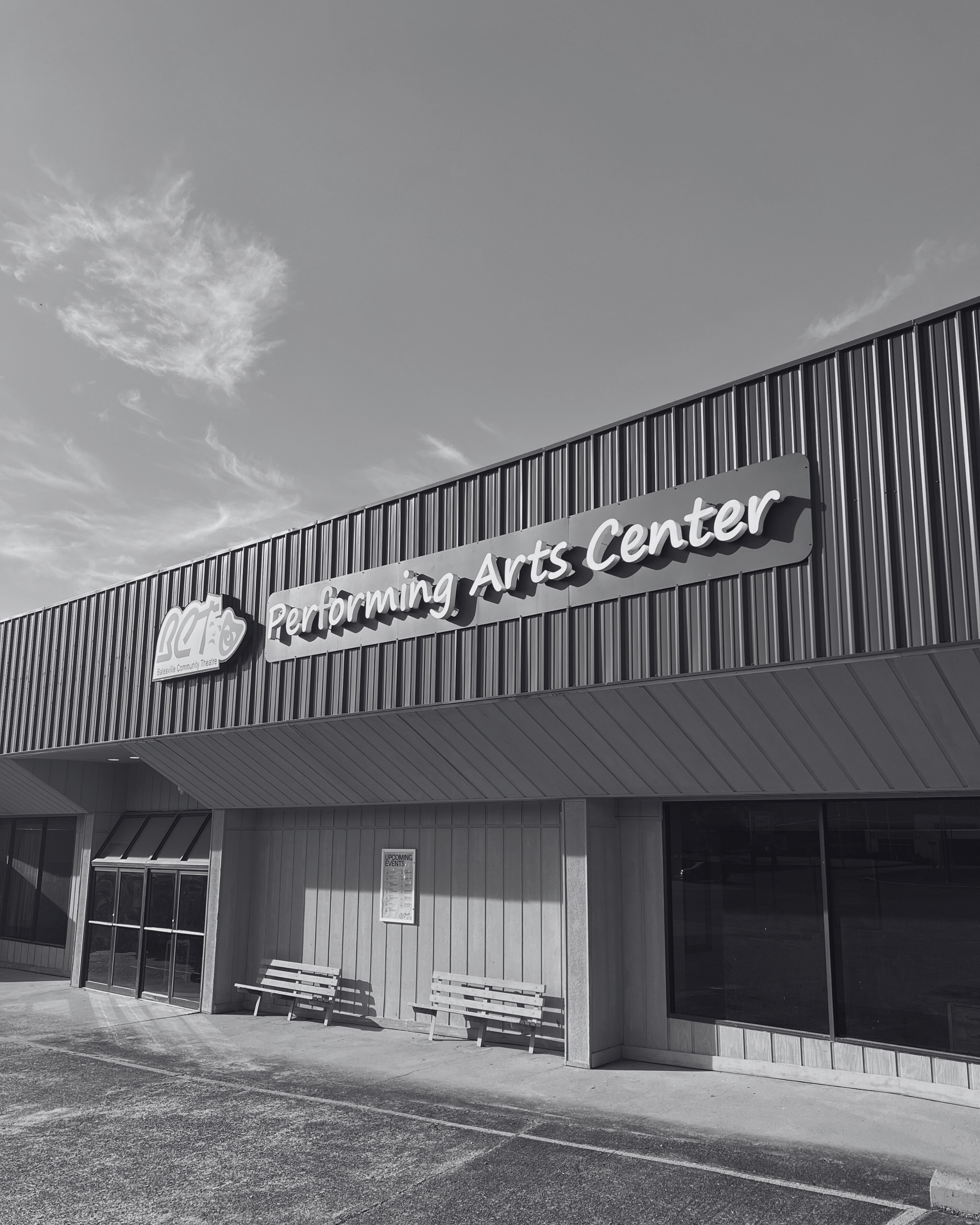 Exterior view of the Performing Arts Center building with a sign, benches outside, and a partly cloudy sky.