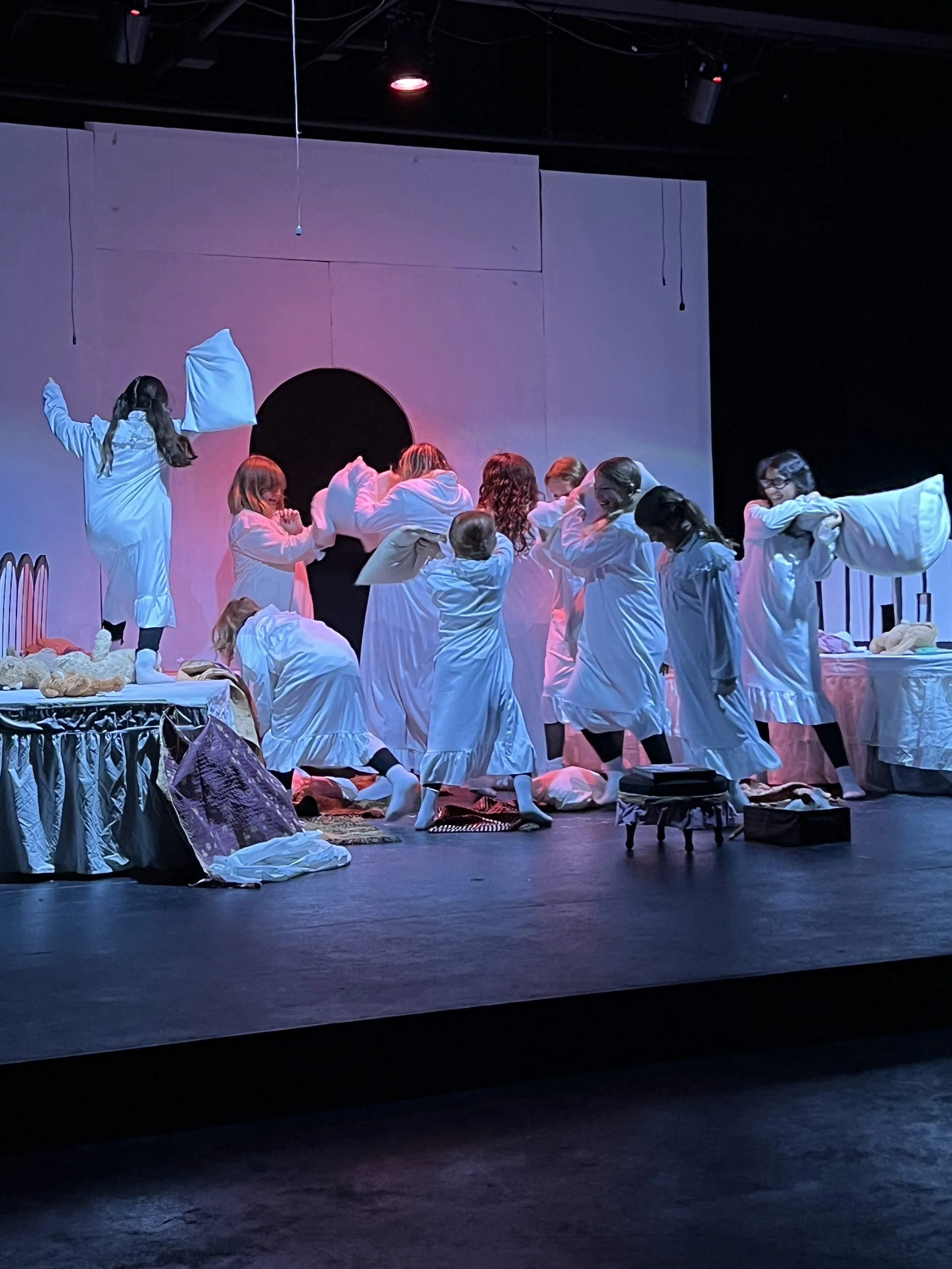 Children in white pajamas engaging in a stage play or performance, with some acting out a scene involving pillows and bed props, in a dimly lit theater with purple and pink lighting.