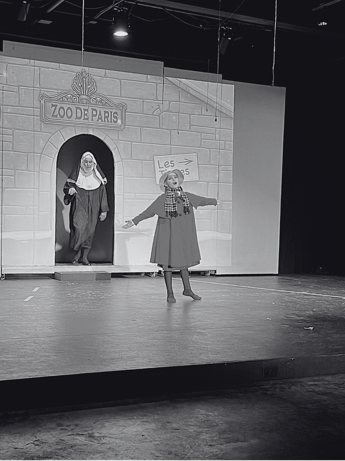 Stage performance with two actors in costume, one in the foreground singing with arms extended, and one in the background emerging from a door of a set labeled 'Zoo de Paris'.