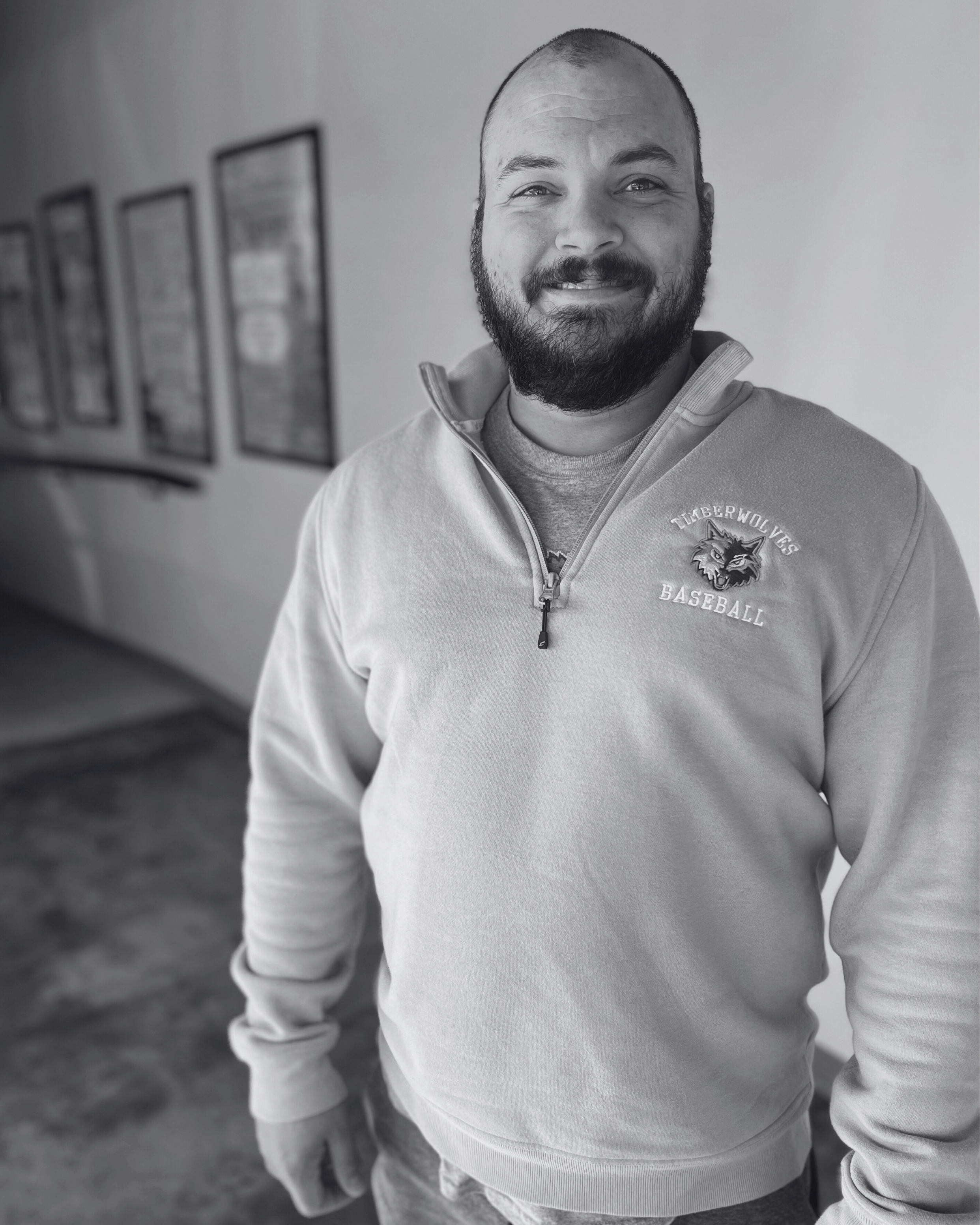 A smiling man with a beard wearing a light blue, quarter-zip sweatshirt with a 'TIGERWOLVES BASEBALL' logo. He stands indoors against a beige wall.