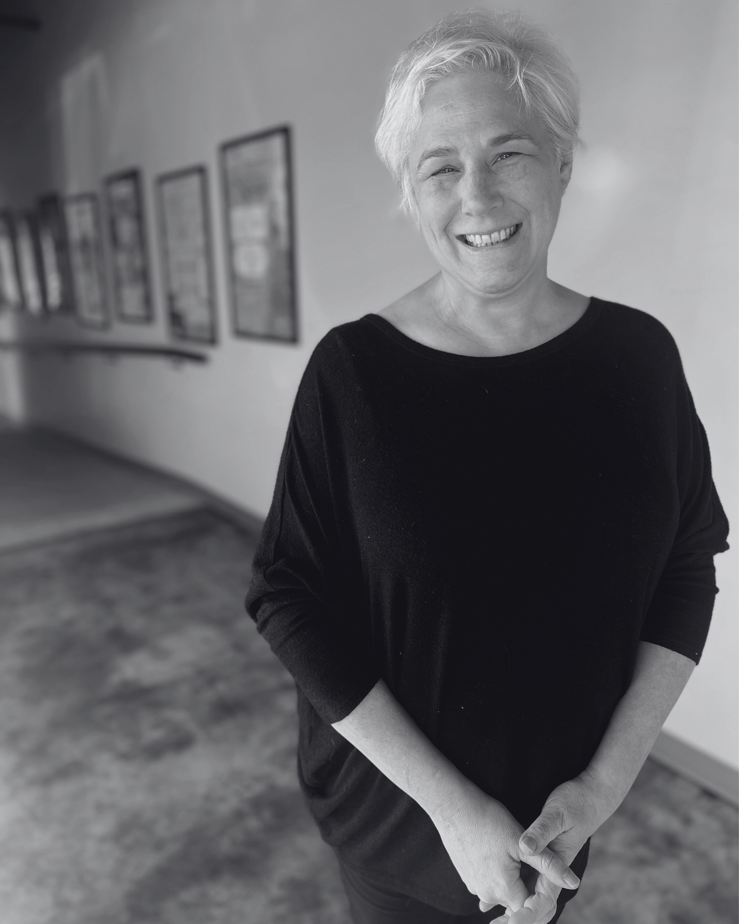 A woman with short blonde hair smiling, wearing a black top, standing in a hallway with framed pictures on the wall.