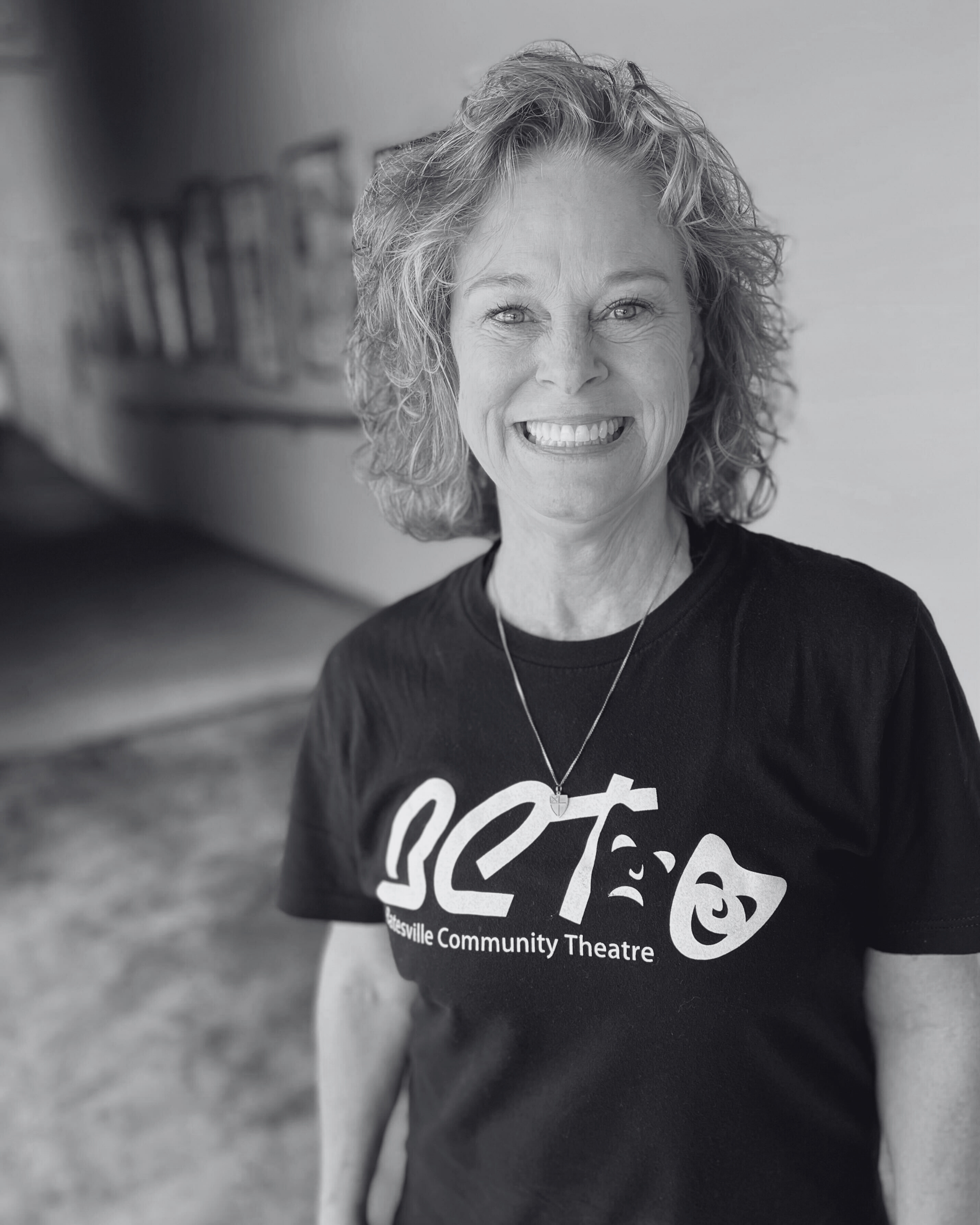 A cheerful woman with curly red hair smiling, wearing a black t-shirt with 'GCT' and 'Gatesville Community Theatre' printed on it, and a gold necklace with a small heart pendant, standing in a hallway with pictures on the wall.