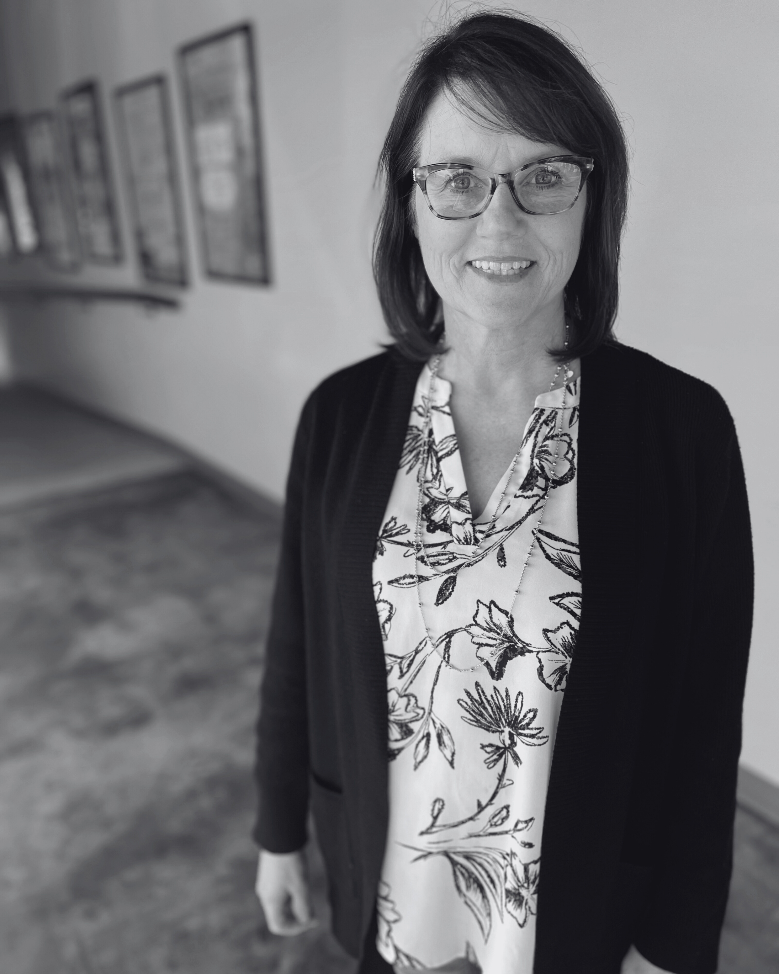 A woman with glasses and brown hair smiling, standing in a hallway with framed pictures on the wall.