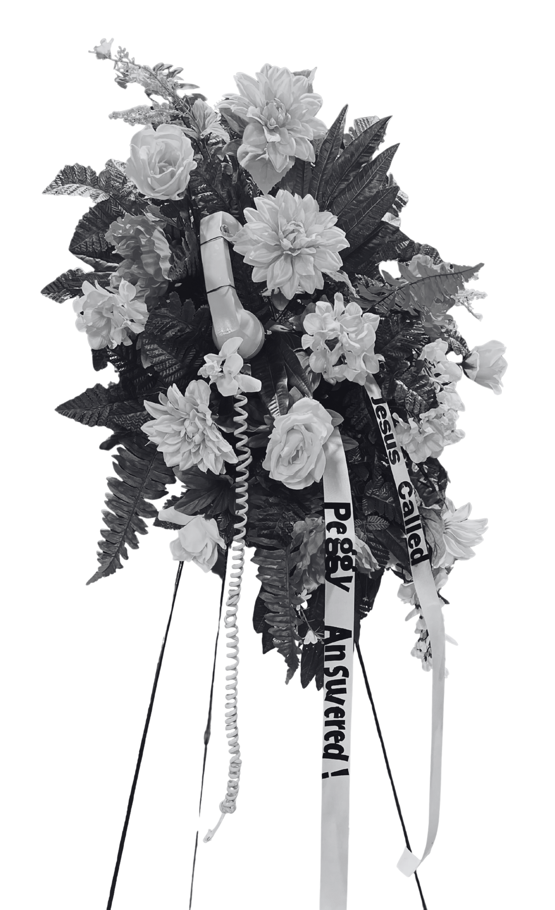 A funeral floral arrangement with roses, dahlias, and greenery, adorned with ribbons that read "Jesus Called," "Pegg," and "As we prayed!" A cordless corded phone is placed among the flowers.