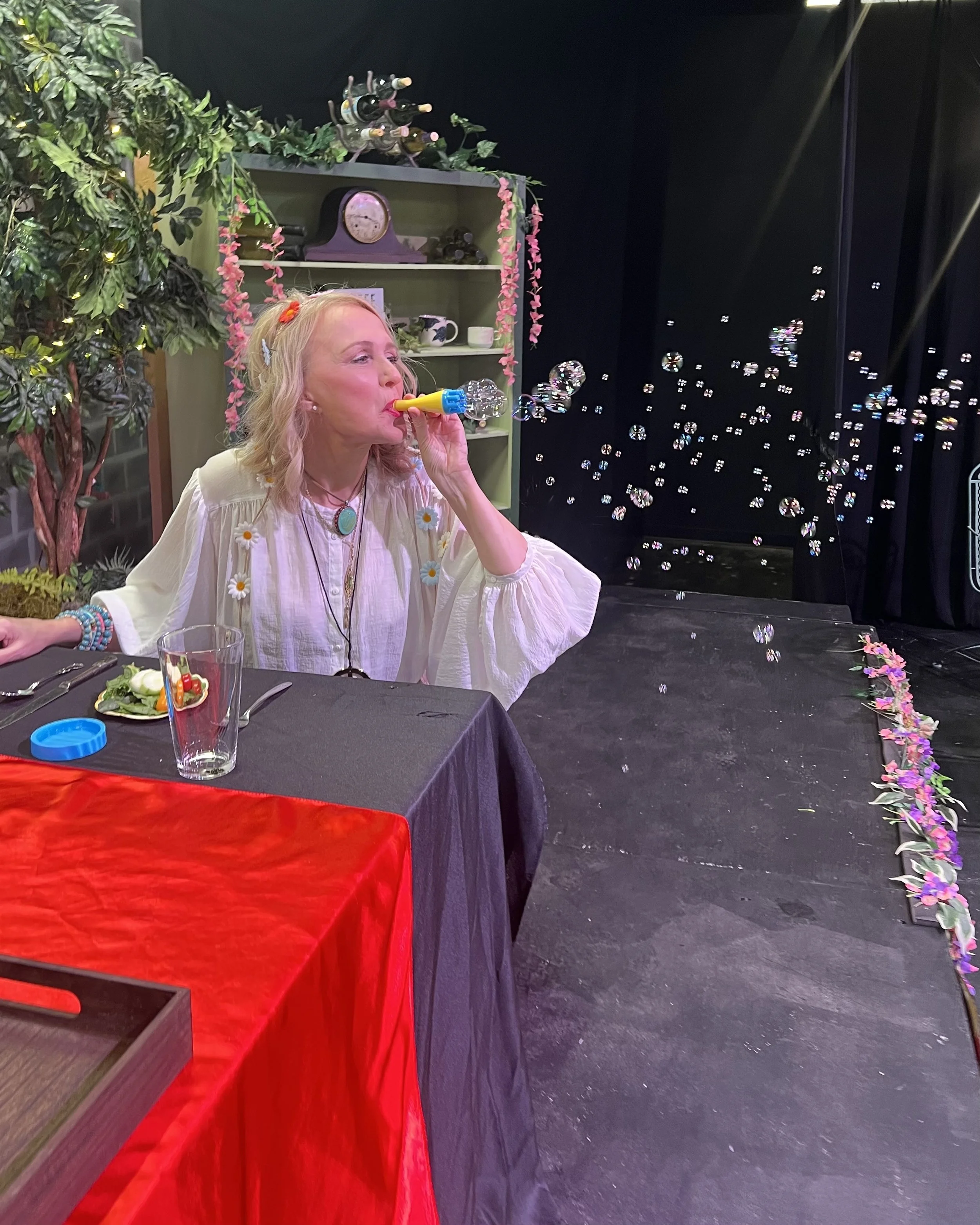 A woman sitting at a table blowing bubbles from a bubble wand in a decorated setting, with a bookshelf and stage curtain in the background.