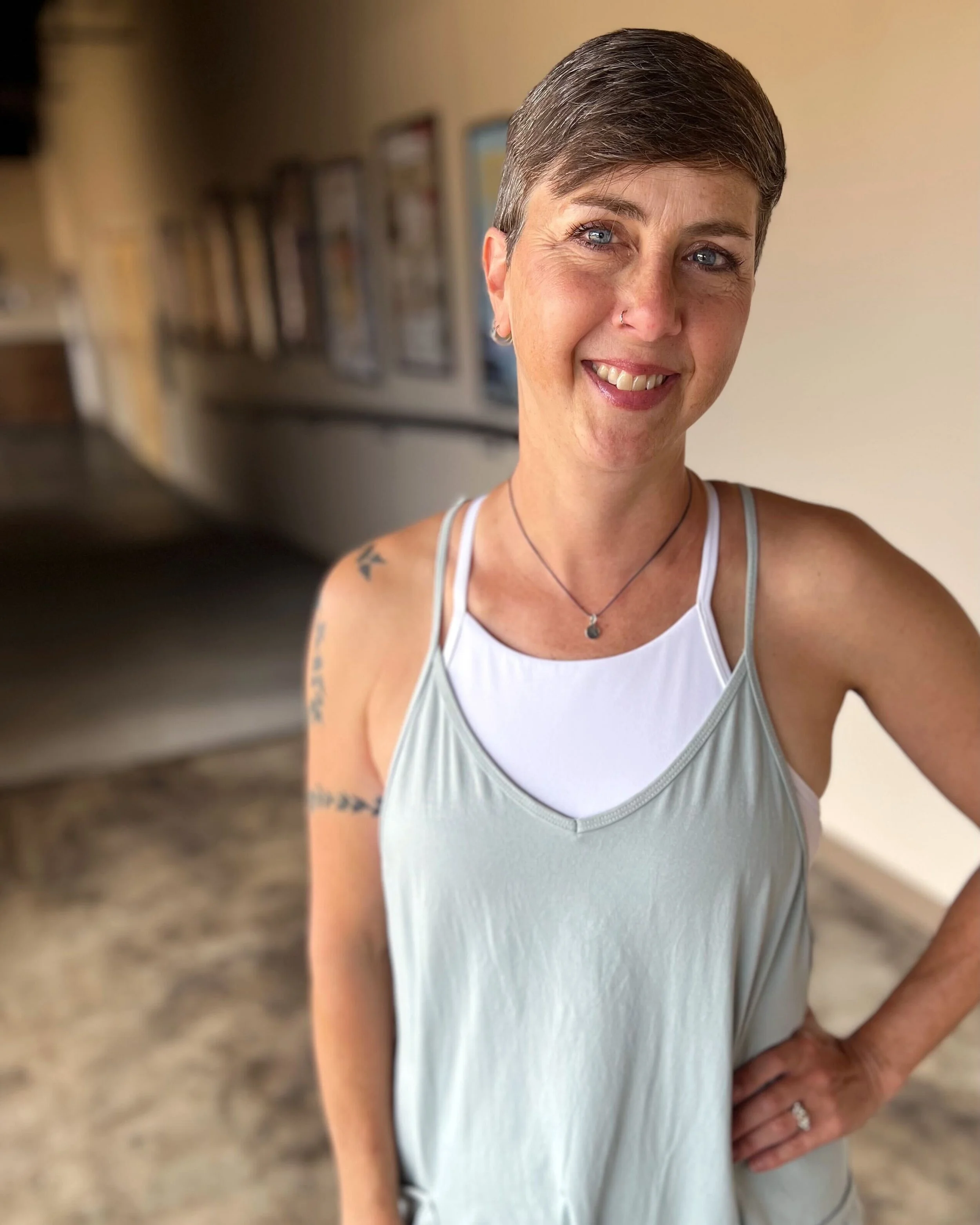 A woman with short hair, blue eyes, and a nose ring taking a selfie. She is wearing a white tank top layered over a white sports bra, a necklace, and has tattoos on her left arm. She is smiling and standing in an indoor setting with blurred artwork on the wall behind her.