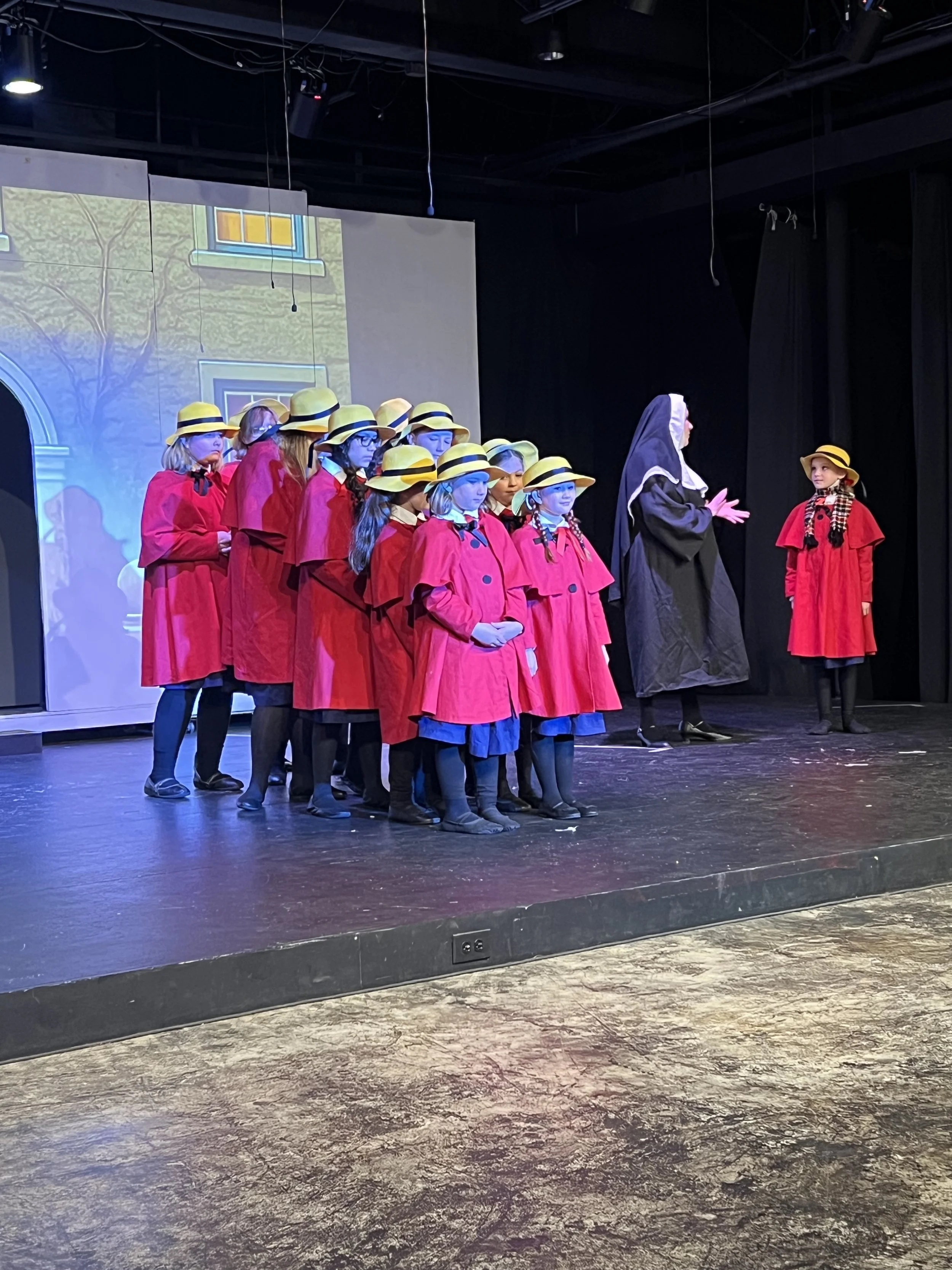 Children in red coats and yellow hats performing on stage with a nun and another girl, with a backdrop of a house and trees.