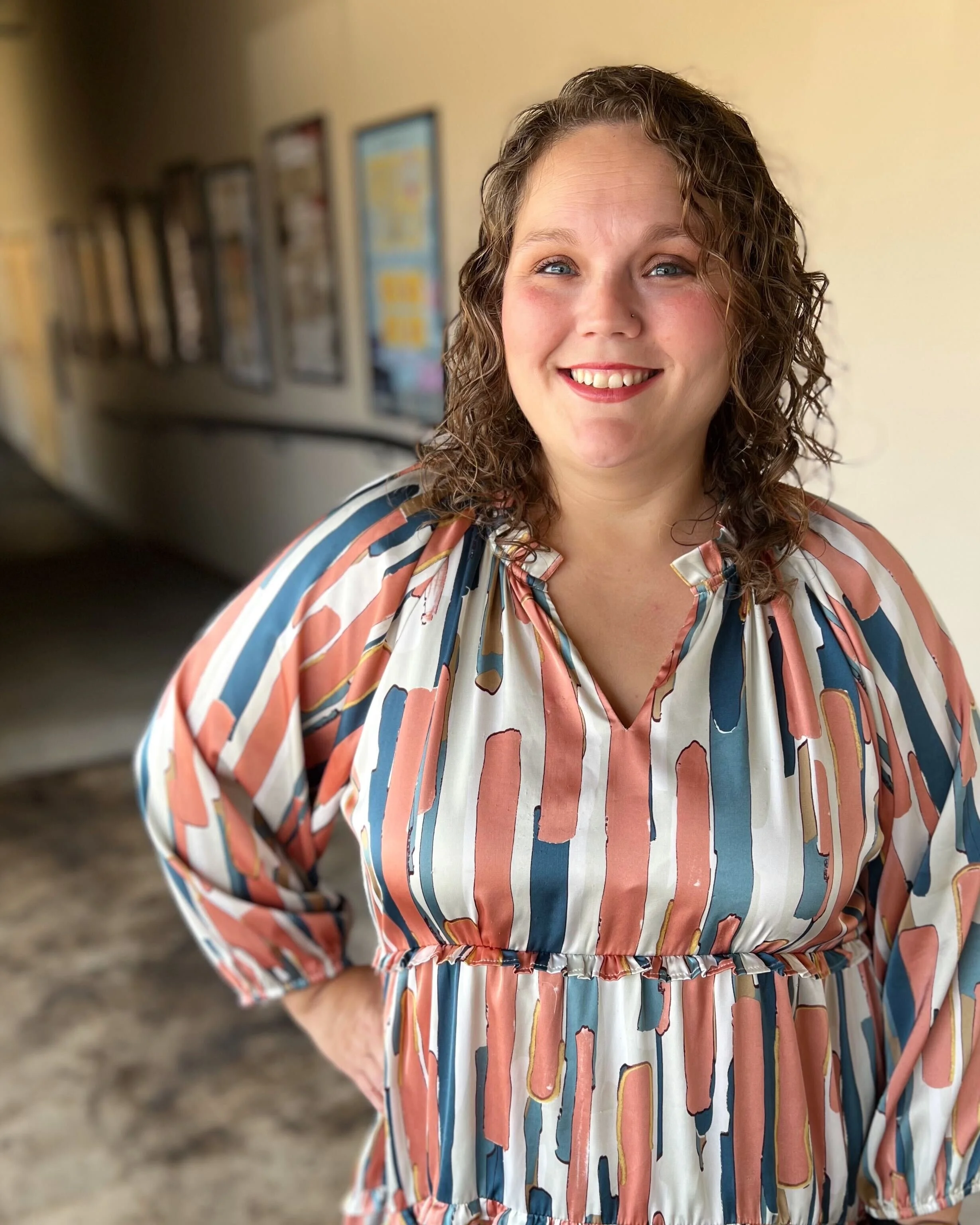 A woman with curly hair smiling, wearing a multicolored striped dress, standing in a hallway with framed photos on the wall.