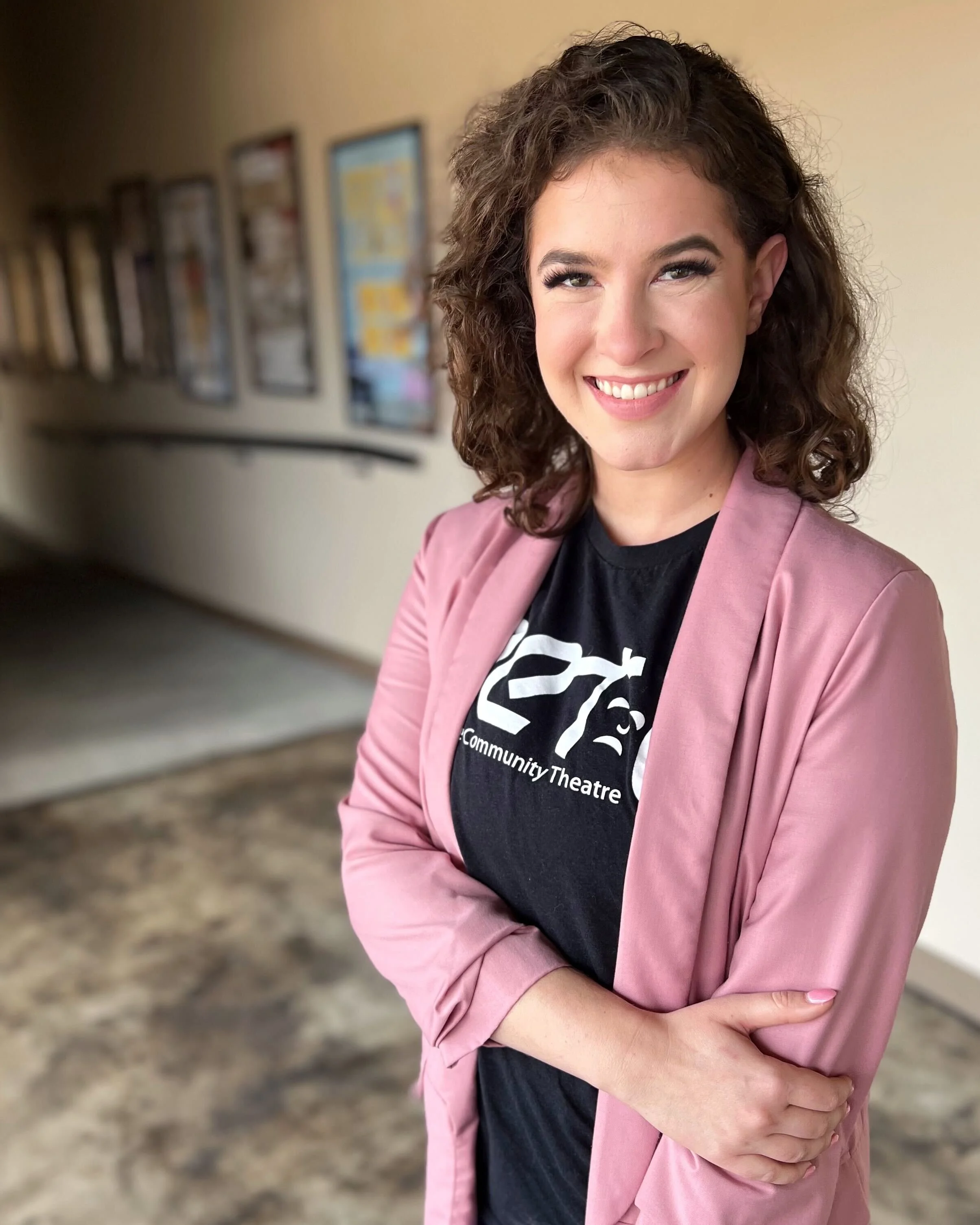 A woman with curly brown hair smiling, wearing a pink blazer and a black T-shirt that reads 'Community Theatre,' standing in a hallway with framed pictures on the wall.