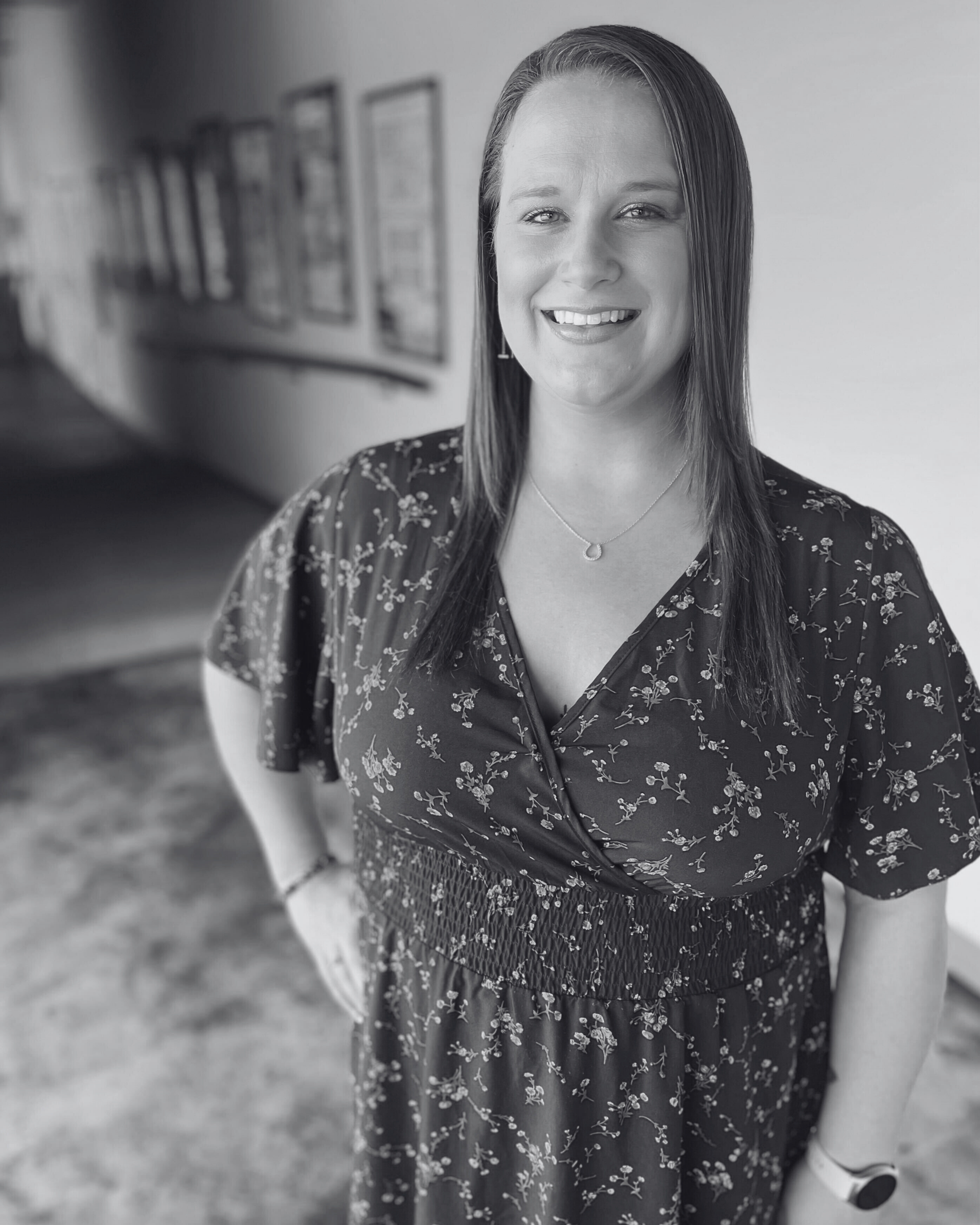 A woman smiling, wearing a blue floral dress, standing in a hallway with framed pictures on the wall.