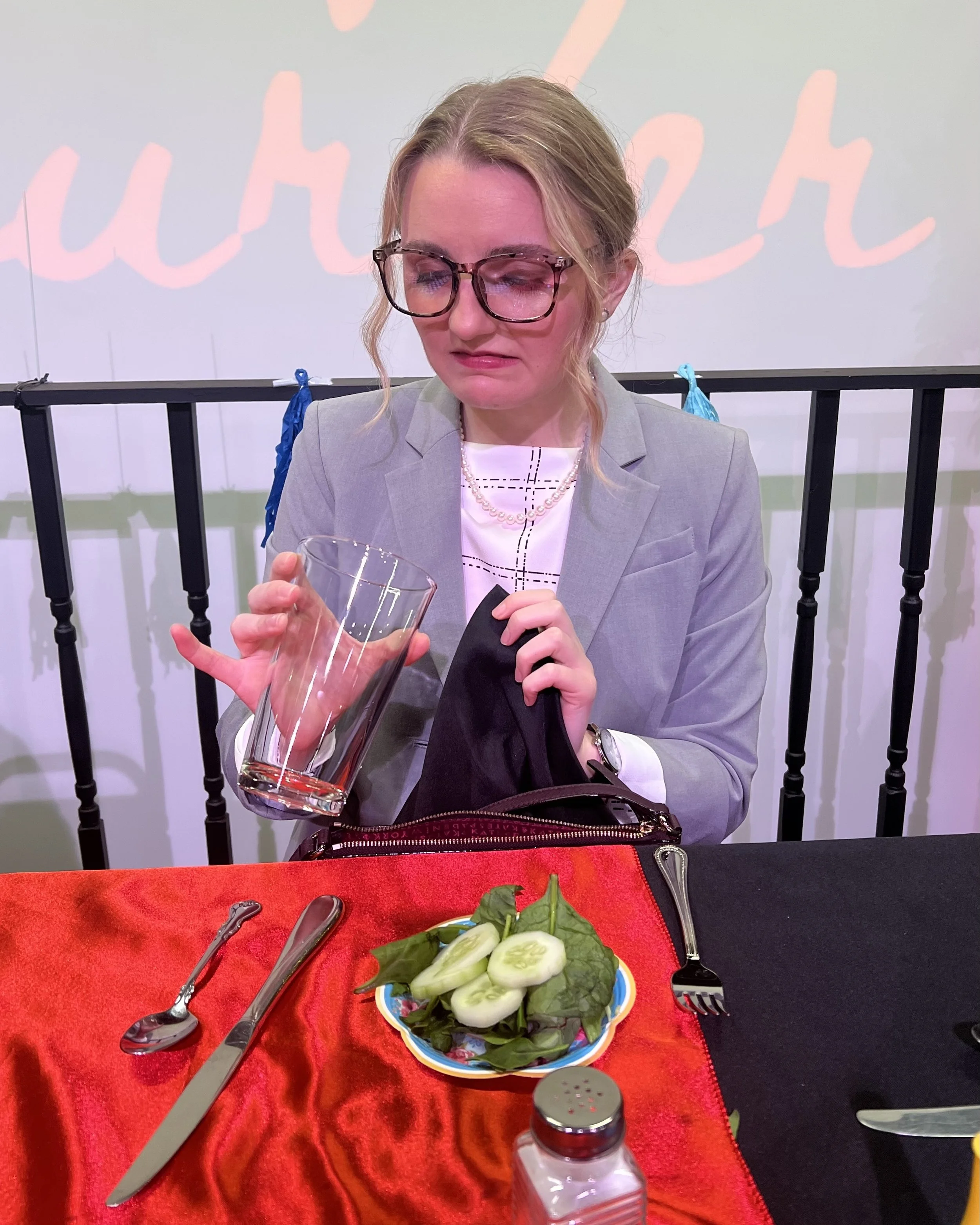 A woman with glasses and a grey blazer looks disappointed while holding an empty glass, sitting at a table with a red tablecloth, a plate of cucumber slices and greens, and utensils.