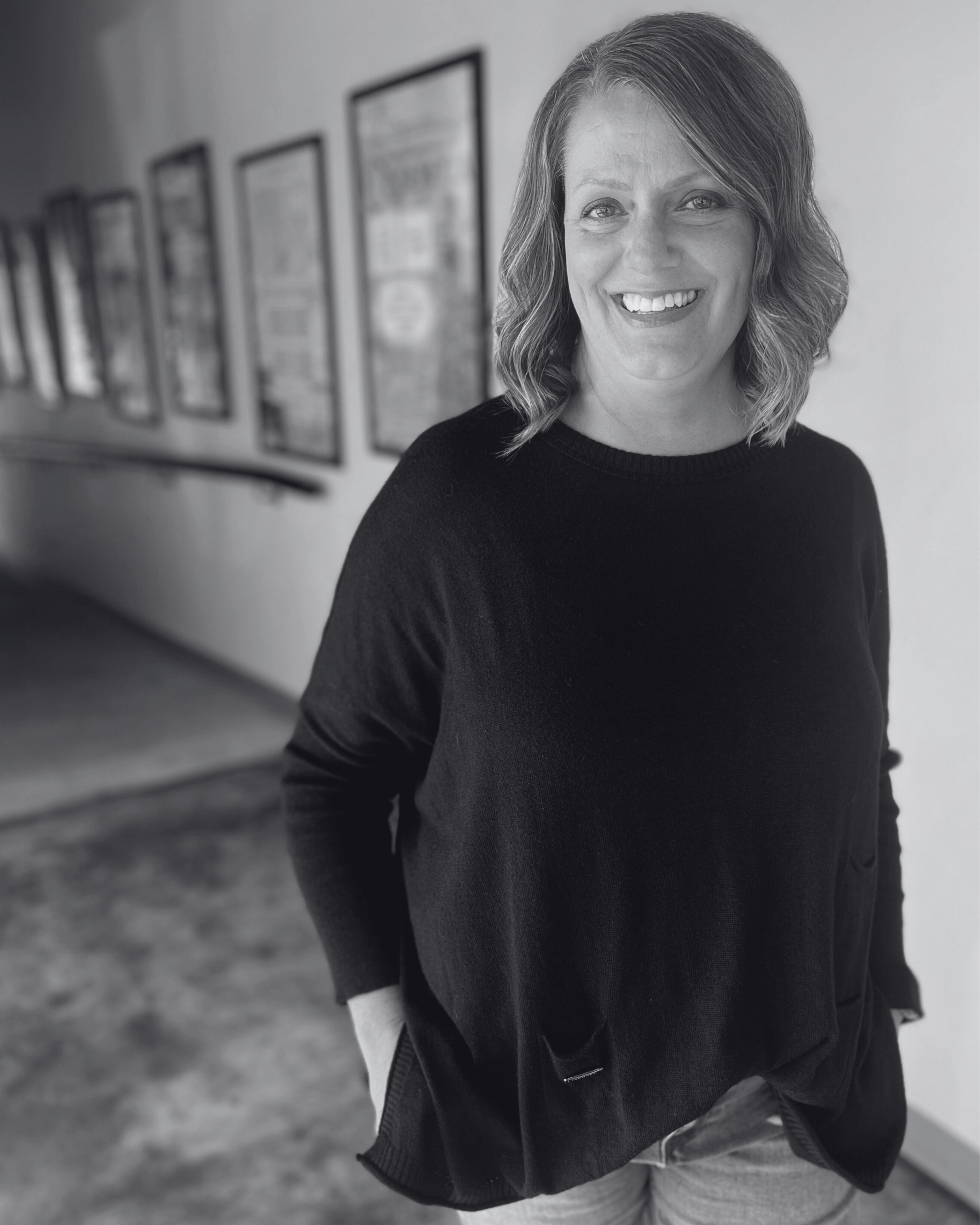 A woman with shoulder-length blond hair, blue eyes, and a wide smile, wearing a black sweater, standing in a hallway with framed pictures on the wall.