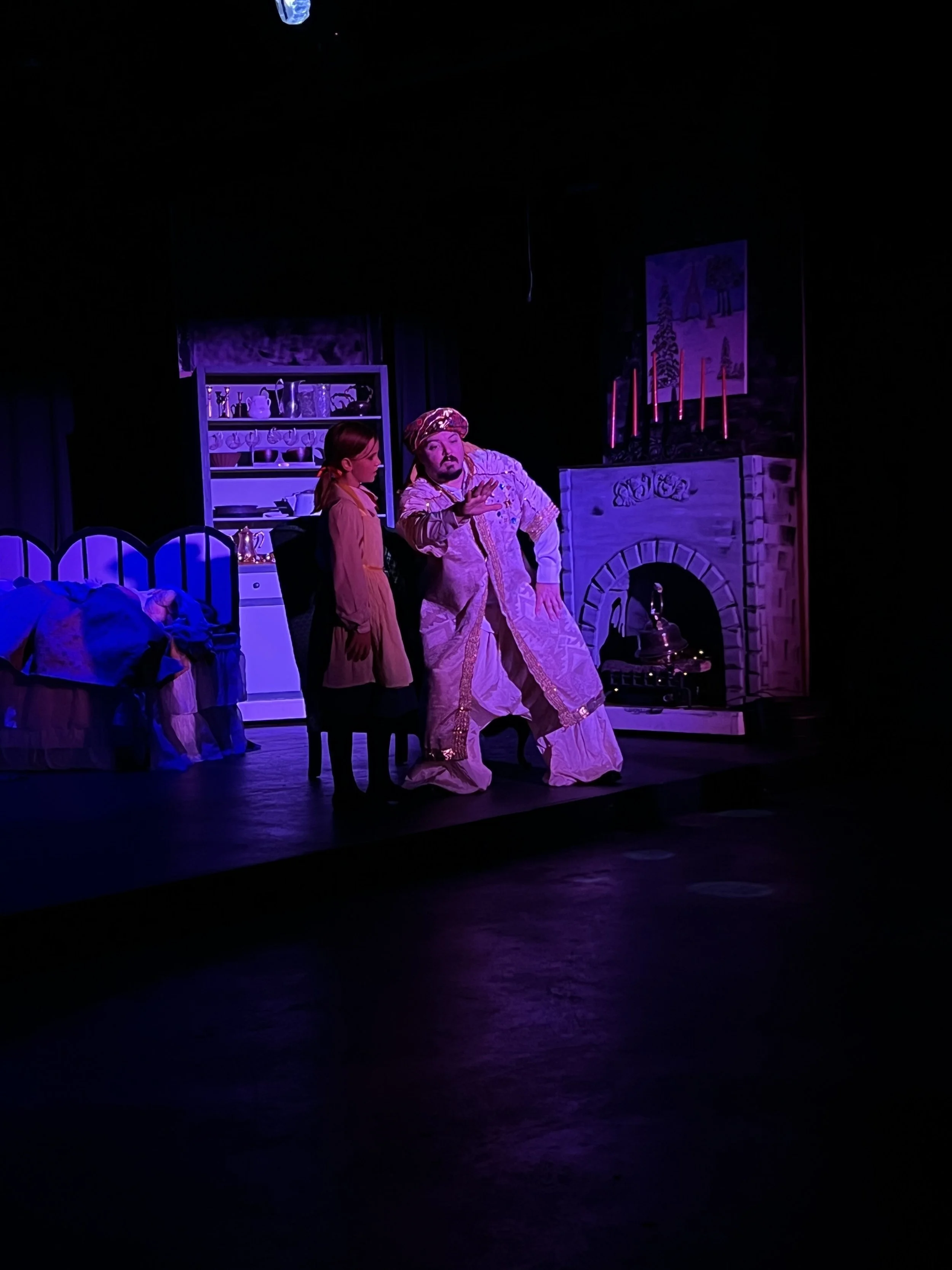 Two actors performing on stage with a fantasy or fairy tale set, including a bed, a shelf, a fireplace, and candles, illuminated with purple lighting.