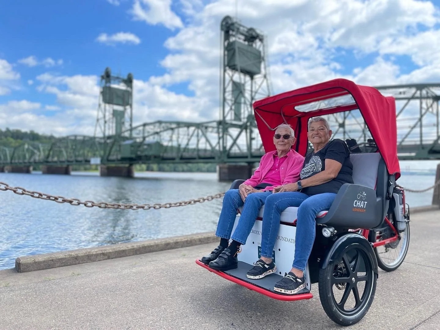 Two older women enjoy a ride in a red adaptive trishaw near the Stillwater Lift Bridge on the St. Croix River. The image highlights accessible outdoor recreation, friendship, and inclusive cycling for older adults and people with mobility challenges.