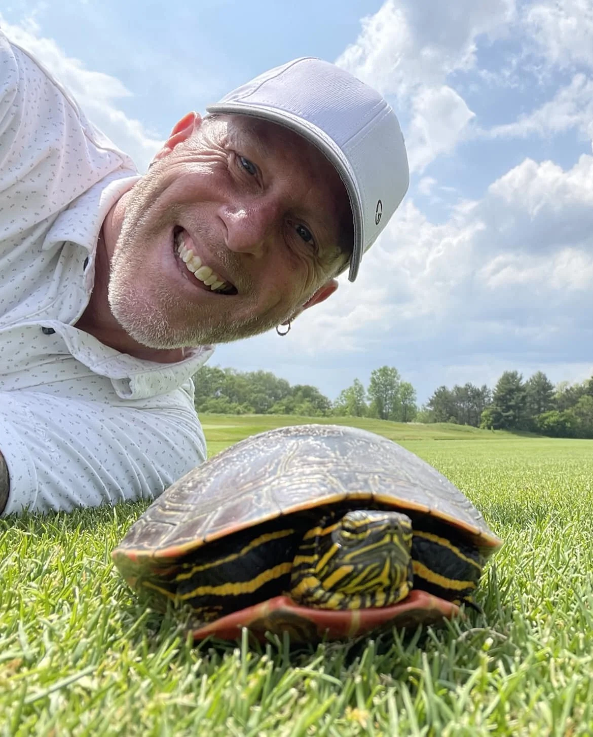 A smiling man lies in the grass beside a painted turtle on a sunny day under blue skies. The image captures a playful moment in nature and reflects Limitless Cycling’s connection to the outdoors, curiosity, and joyful community experiences.
