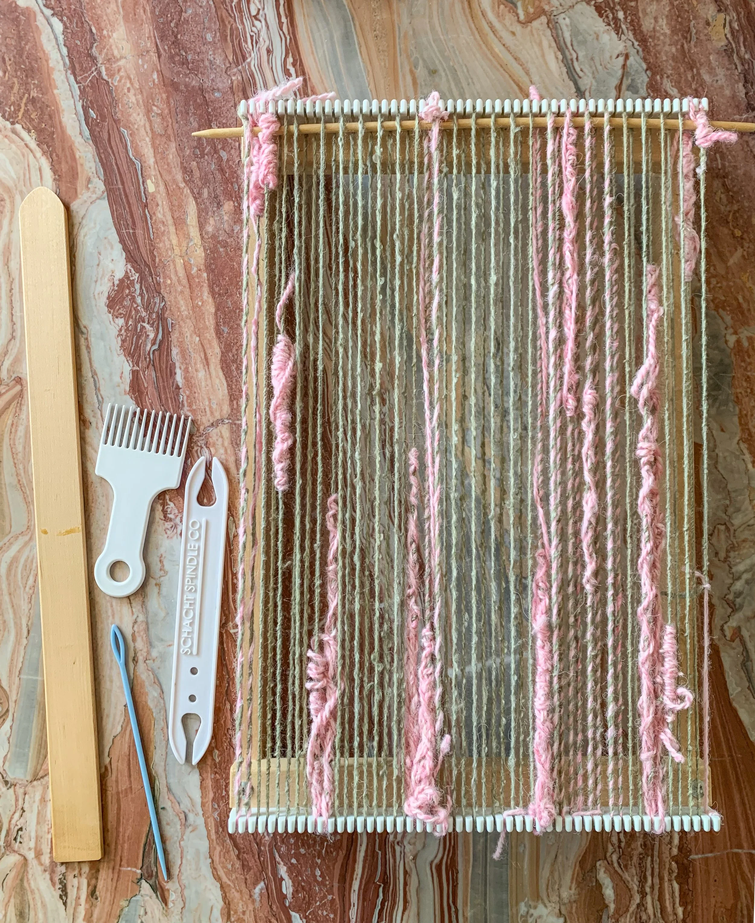 A small weaving loom with pink and beige yarns, a wooden beater, and a yarn pointer placed on a marble surface.