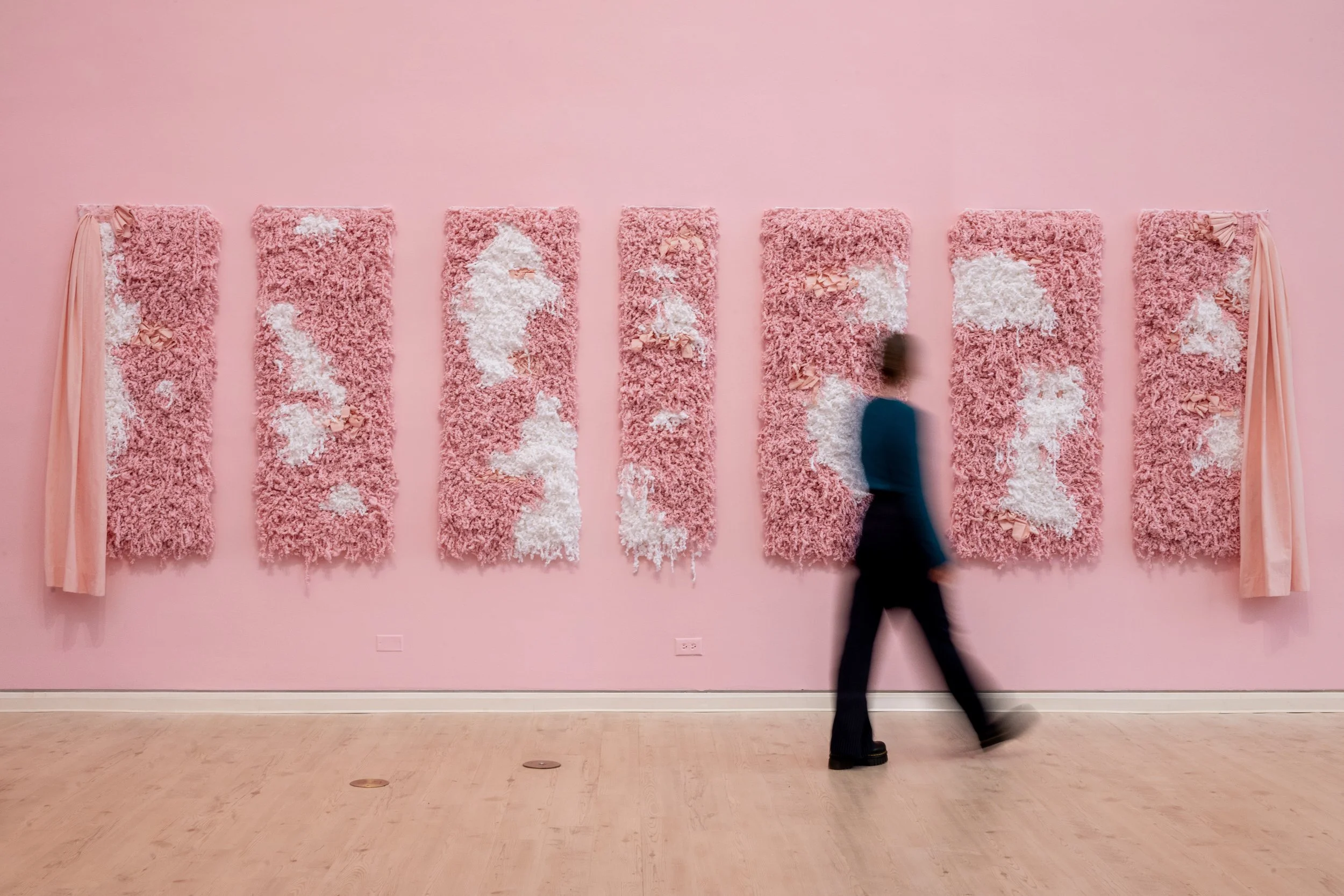 A woman wearing black walking to the left of a pink and white weaving installation named Priscilla Made