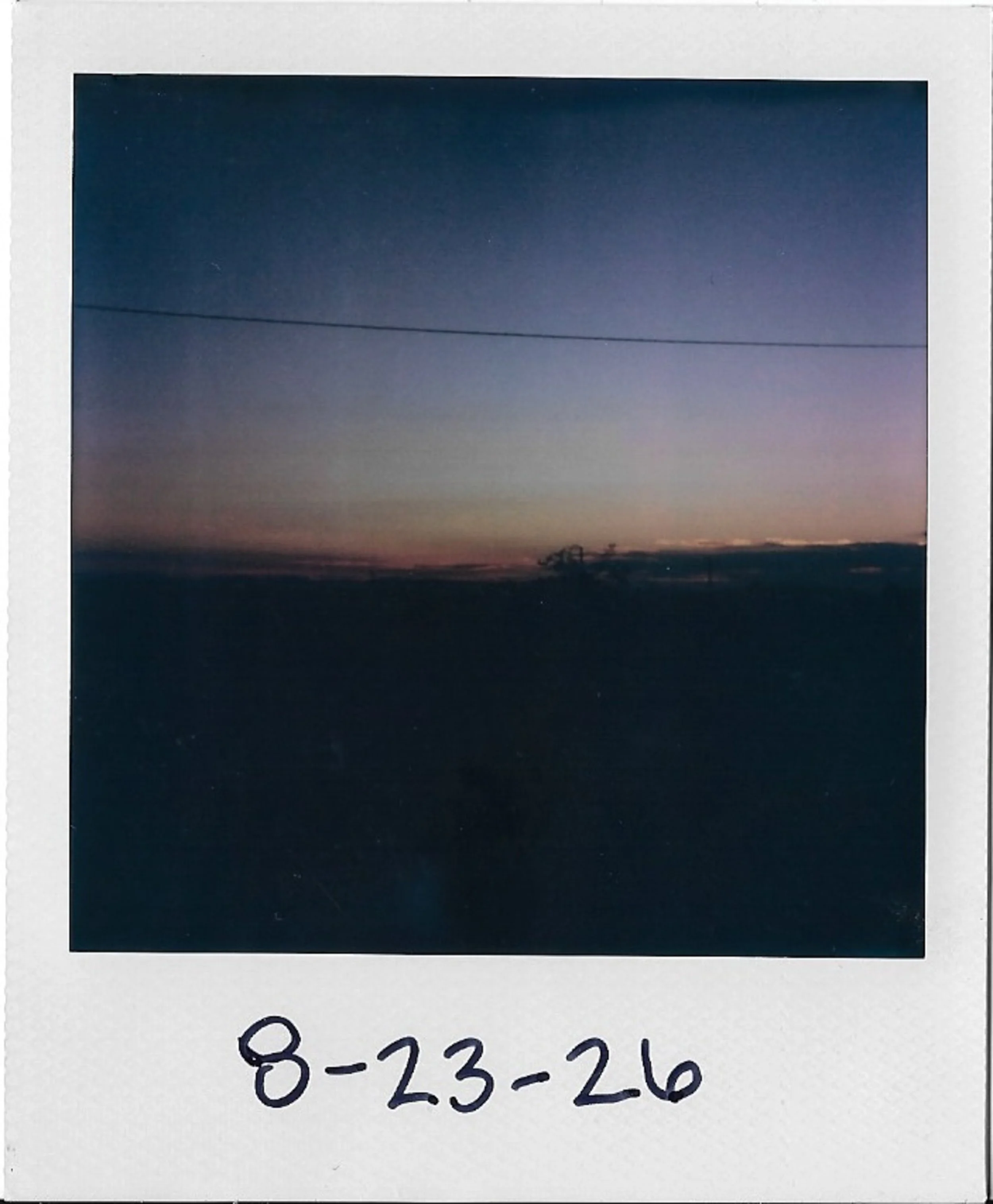 A Polaroid photograph showing a sunset sky with clouds, a silhouette of trees, and a power line against the horizon.