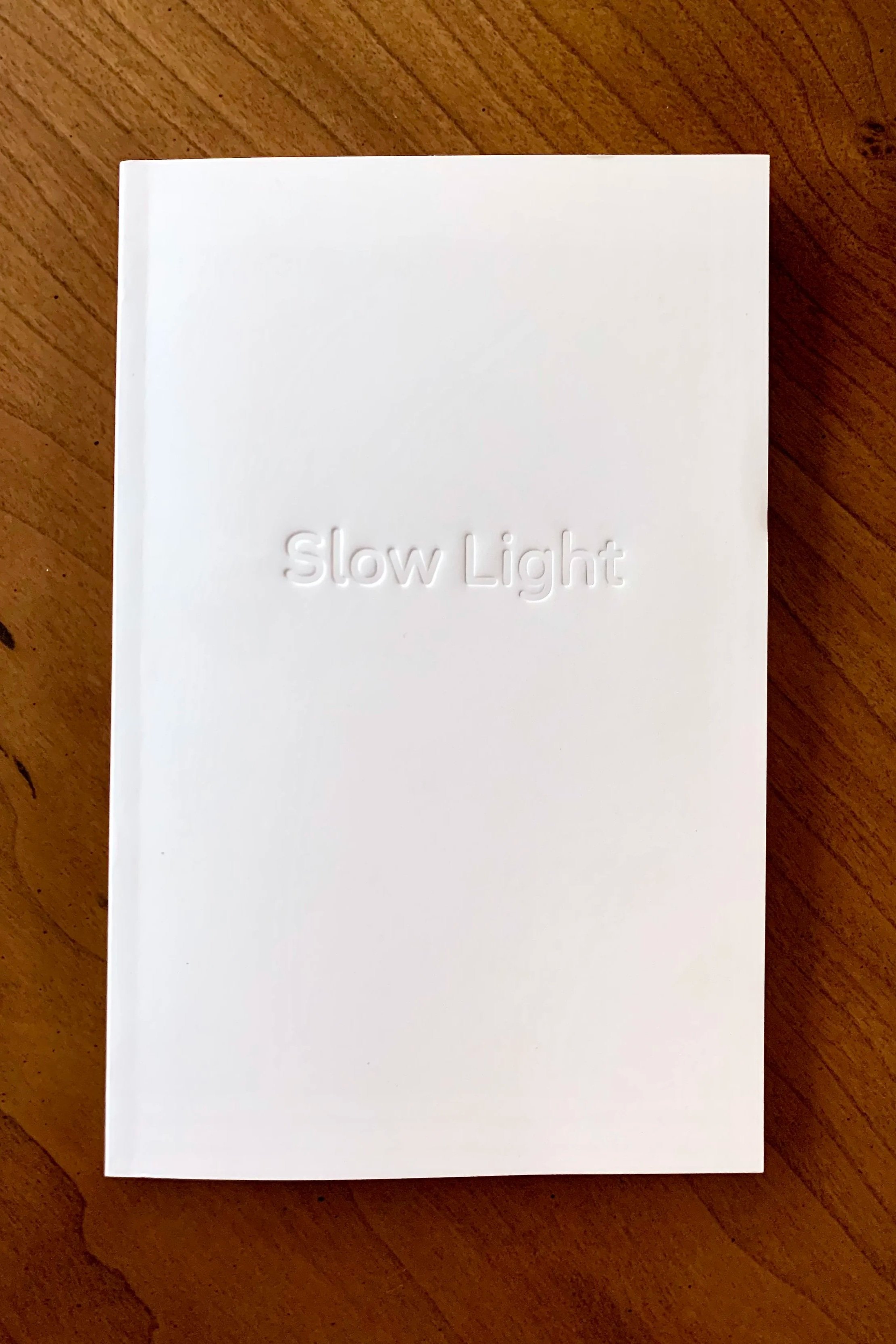 A white booklet or card with the embossed words 'Slow Light' on the cover, placed on a wooden surface.