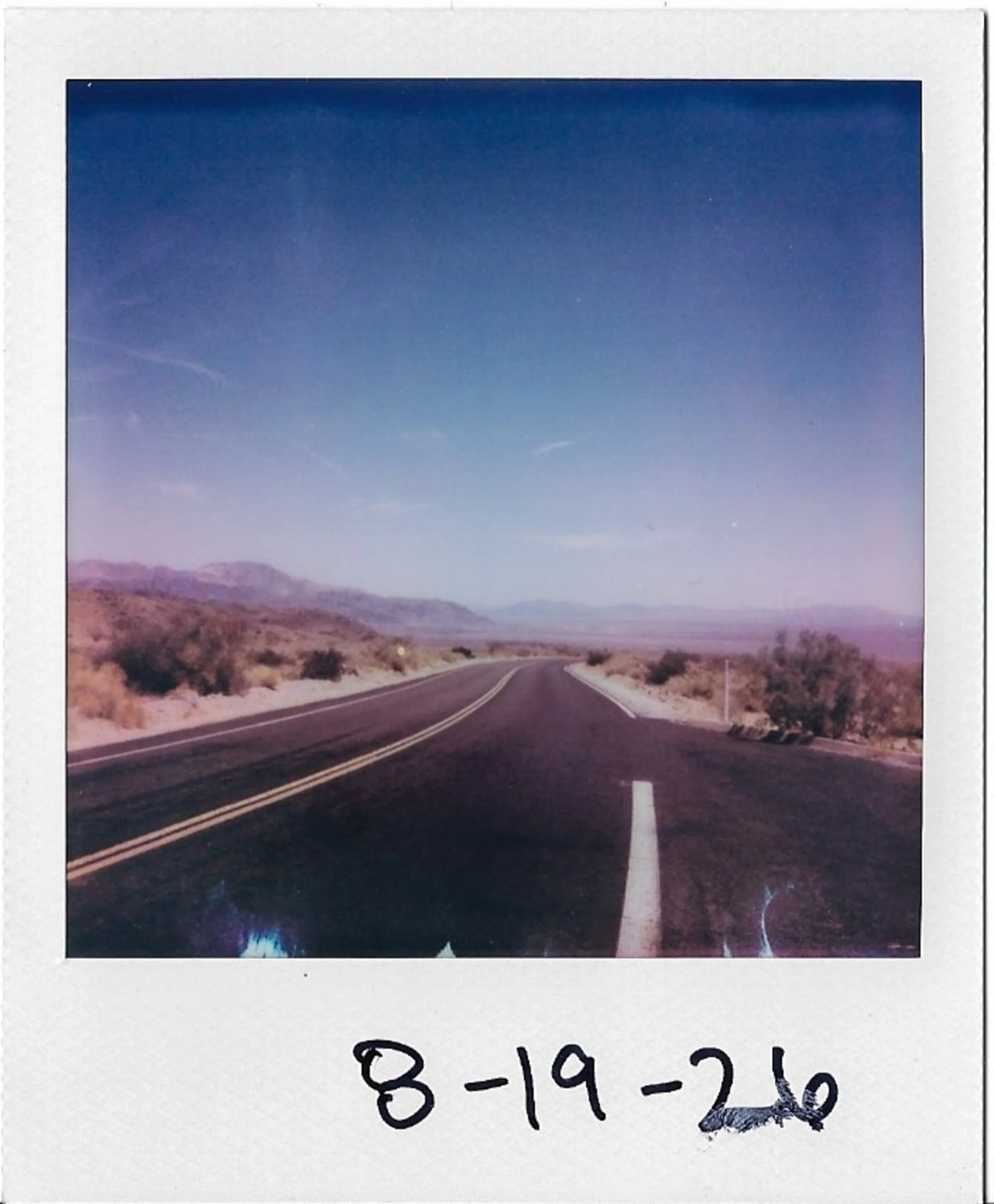 Photograph of a polaroid picture showing a desert landscape with mountains in the distance and a road in the foreground, under a clear blue sky, with handwritten date '8-19-20' at the bottom.