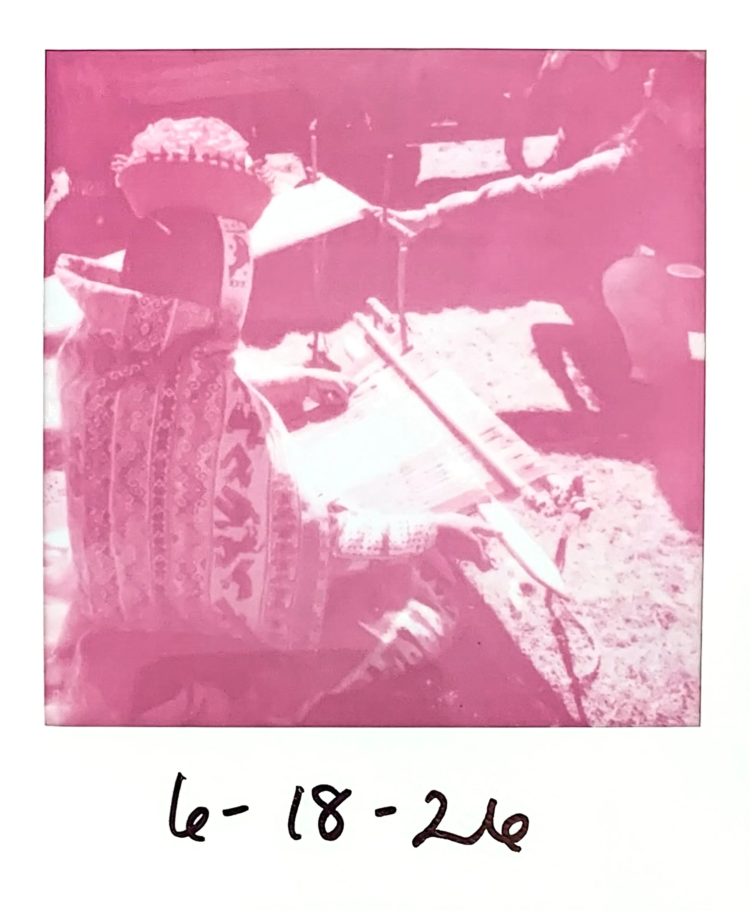 A pink-tinted photograph of a Quechua weaver demonstrating how to weave on a backstrap loom. The photograph is dated '6-18-26'.