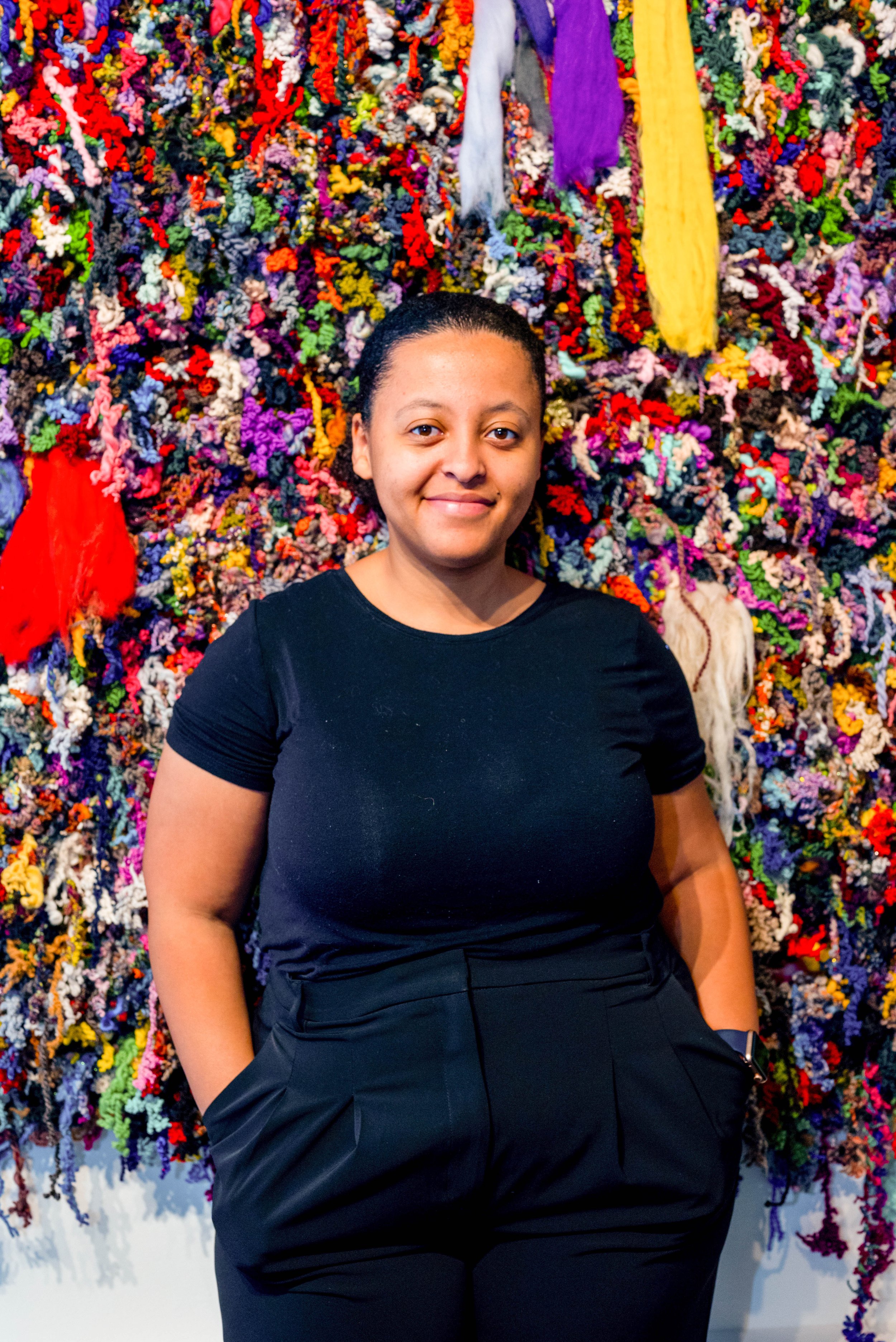 A woman with short black hair, wearing a black T-shirt and black pants, stands in front of a colorful, textured textile artwork background.