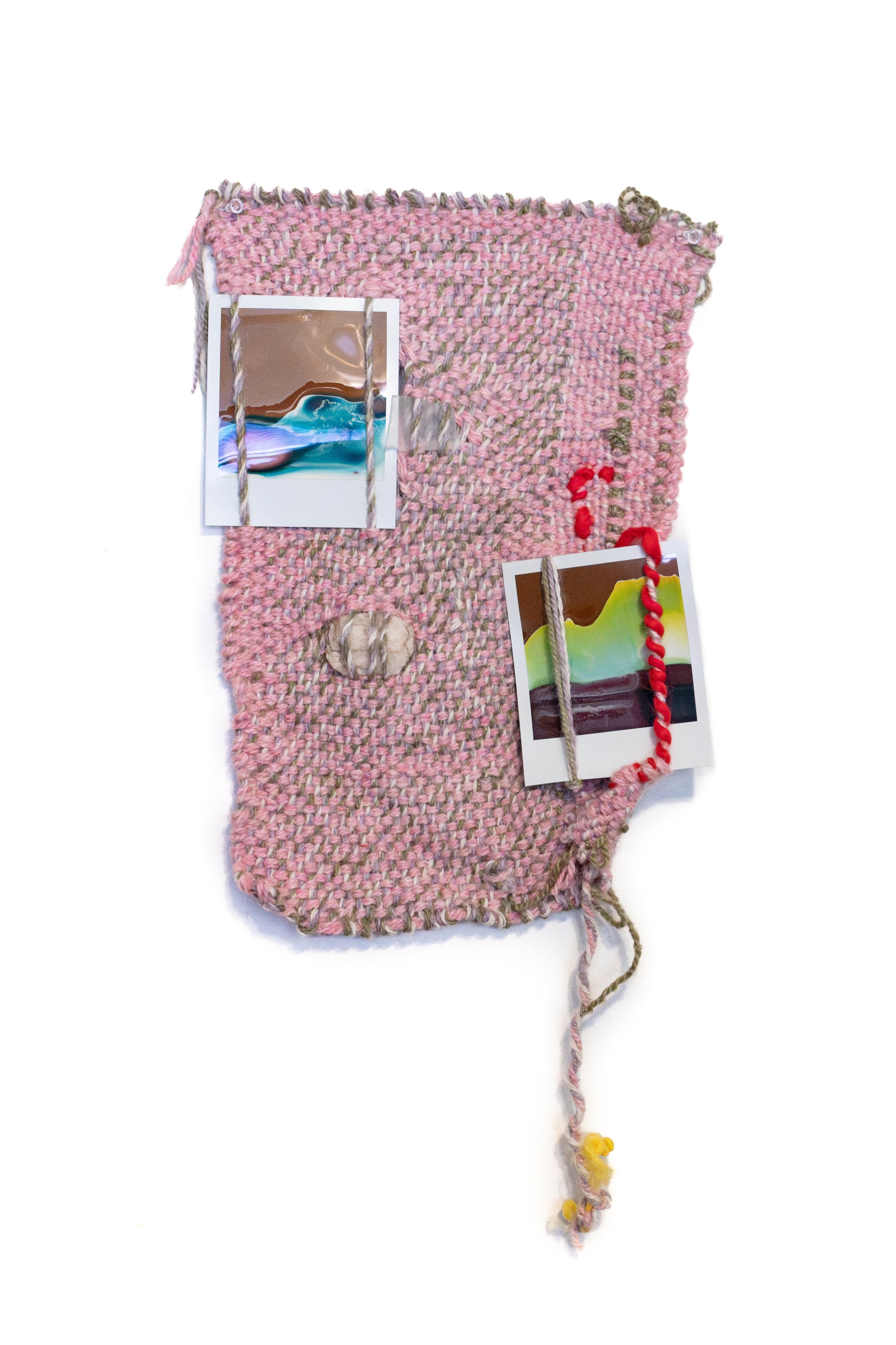 A pink woven textile with various small decorative items attached, including two failed multicolored polaroids and a rock.