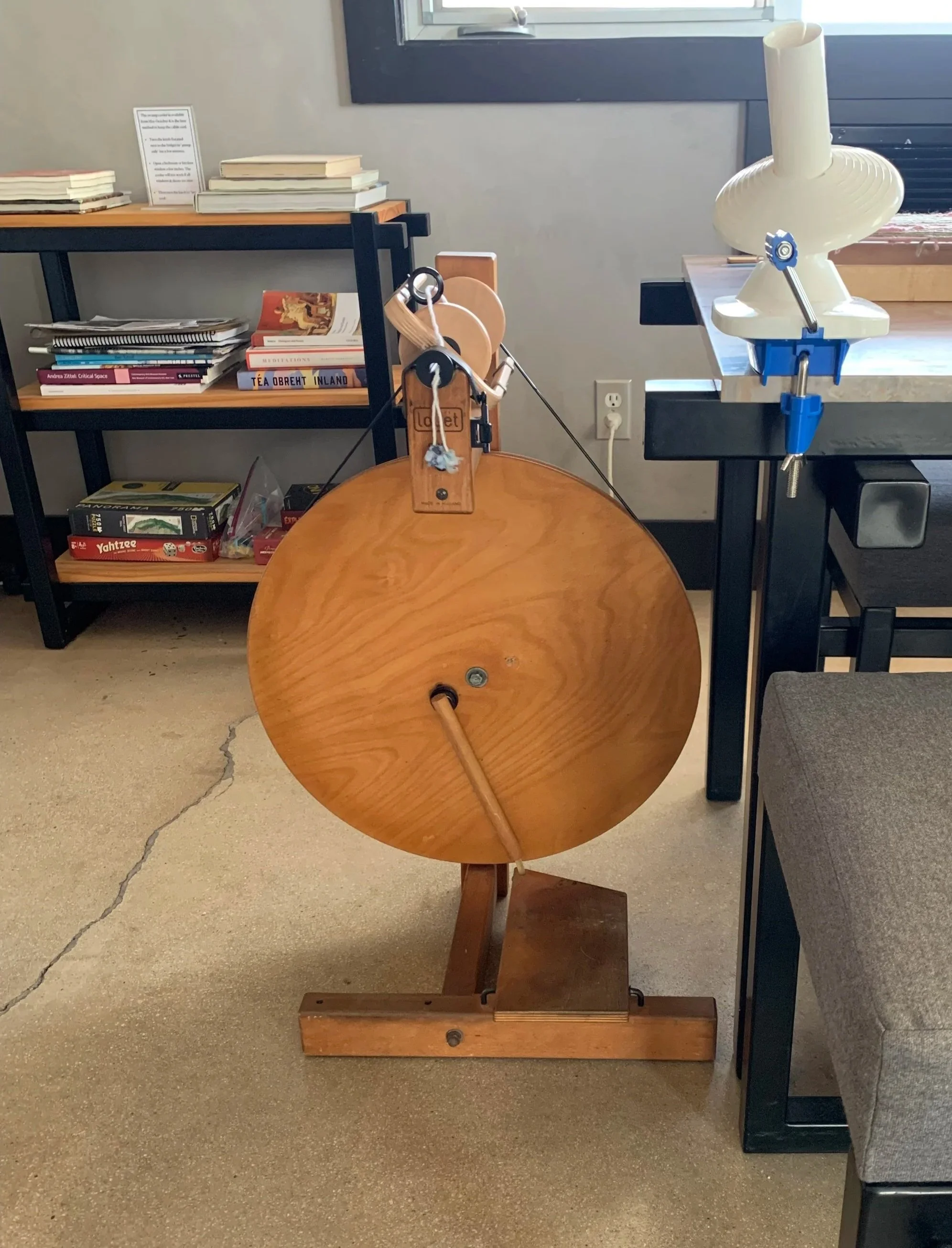 A wooden spinning wheel and a table in a room with bookshelves and a window.