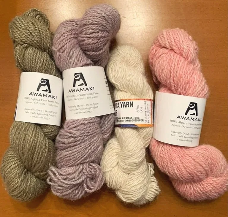 Four skeins of yarn in different colors: olive green, lavender, off-white, and pink, placed on a wooden surface.