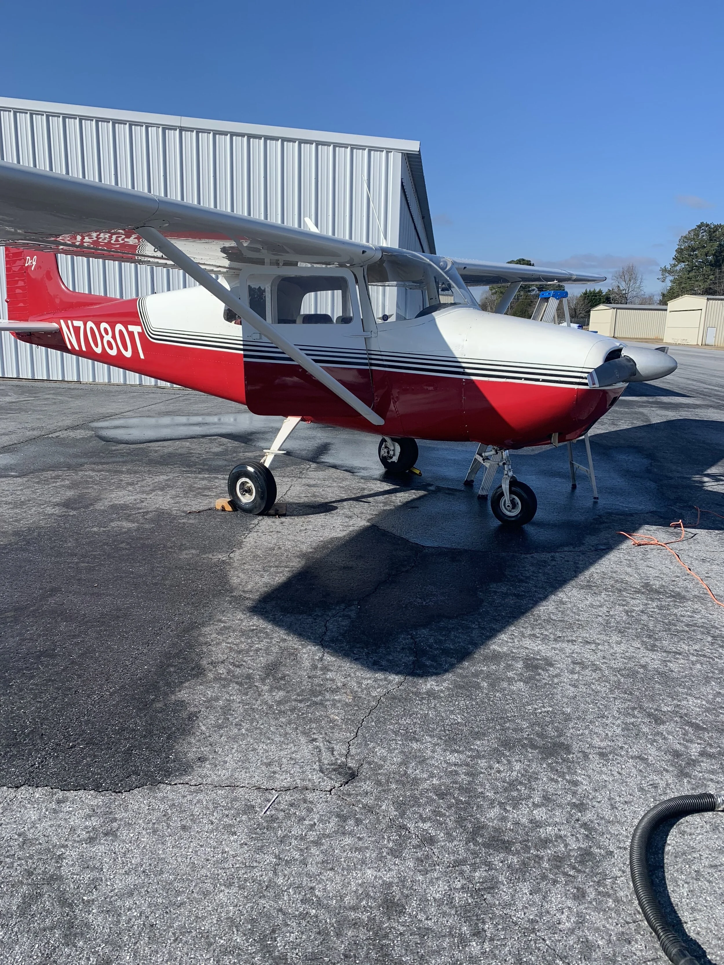 Small red and white Cessna airplane parked on tarmac outside hangar with blue sky and trees in background.