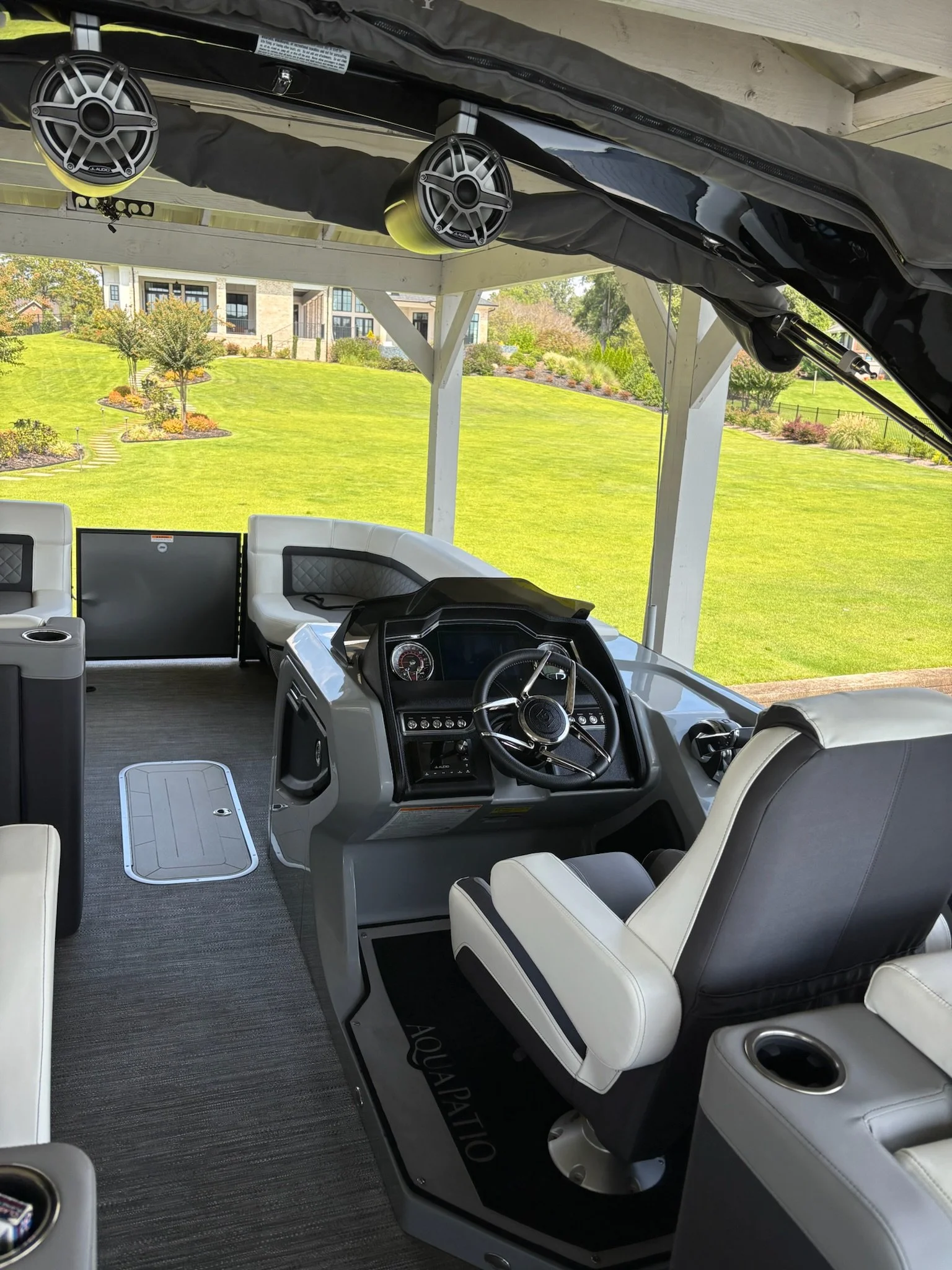 Interior of a luxury pontoon boat with captain's seat, steering wheel, dashboard, and seating area, overlooking a green lawn and houses outside.