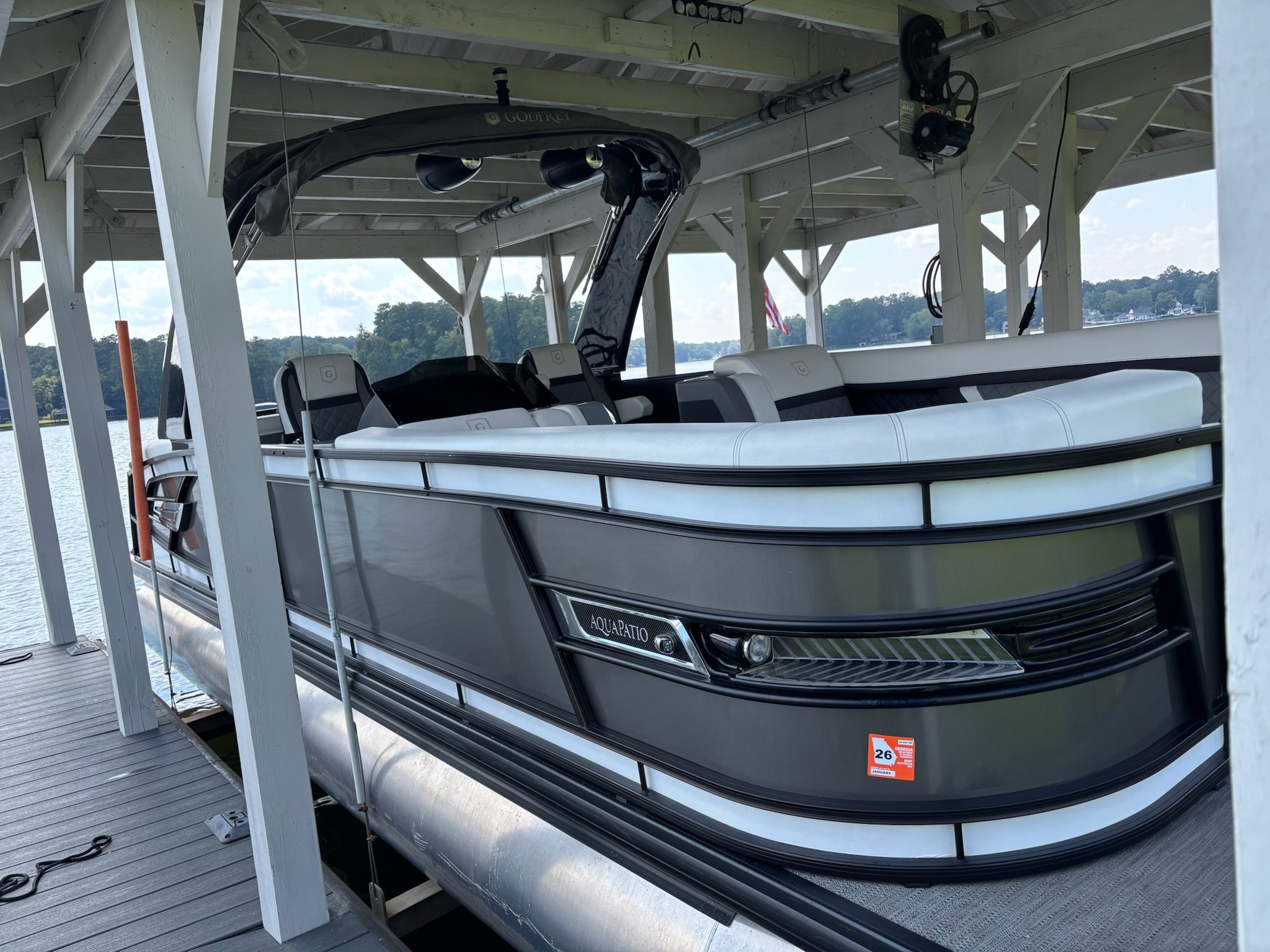 A modern gray pontoon boat docked at a marina on a lake, with a lake and trees visible in the background.