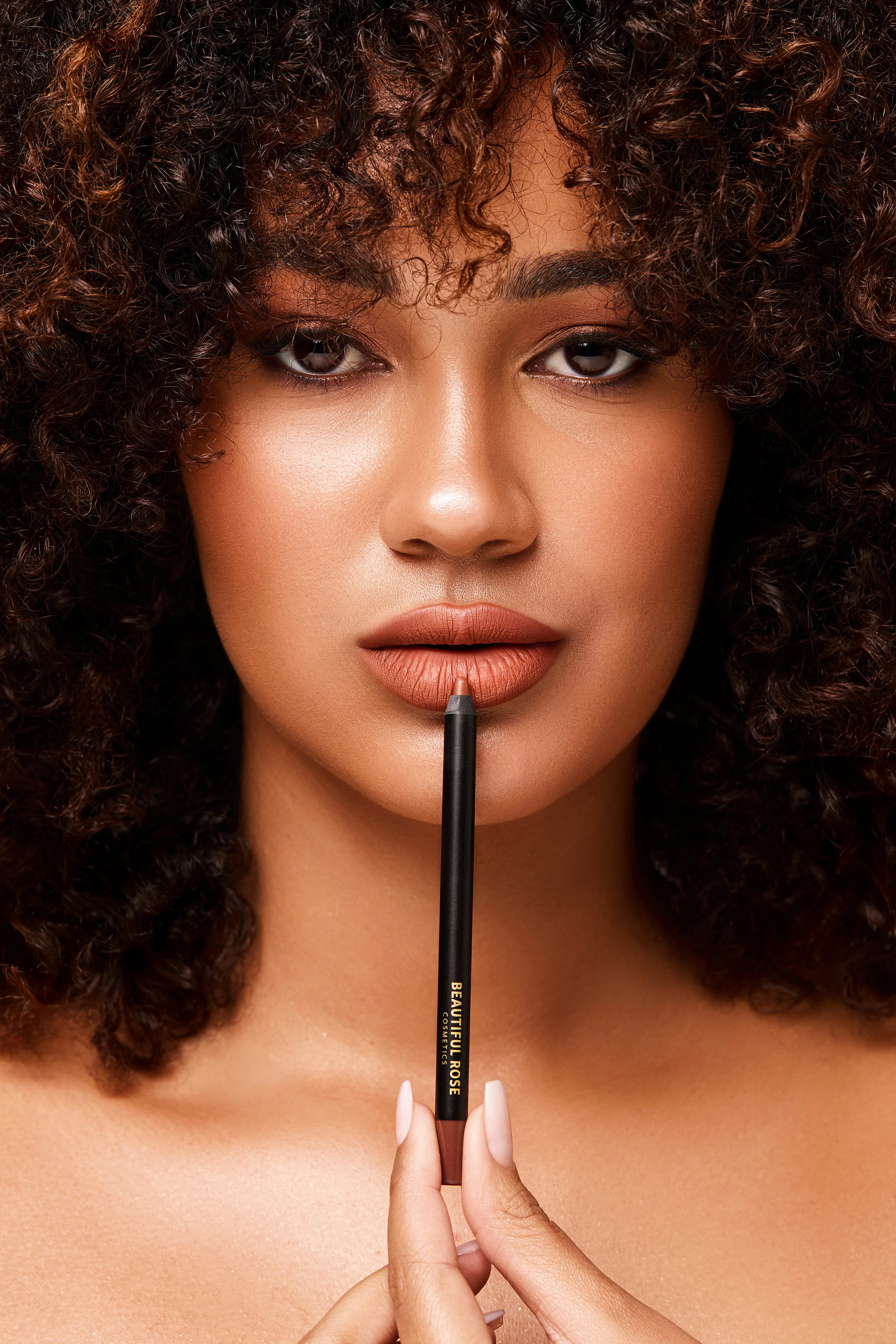 Close-up of a woman with curly hair holding a lip brush to her lips, which are painted with nude lipstick, with a neutral background.