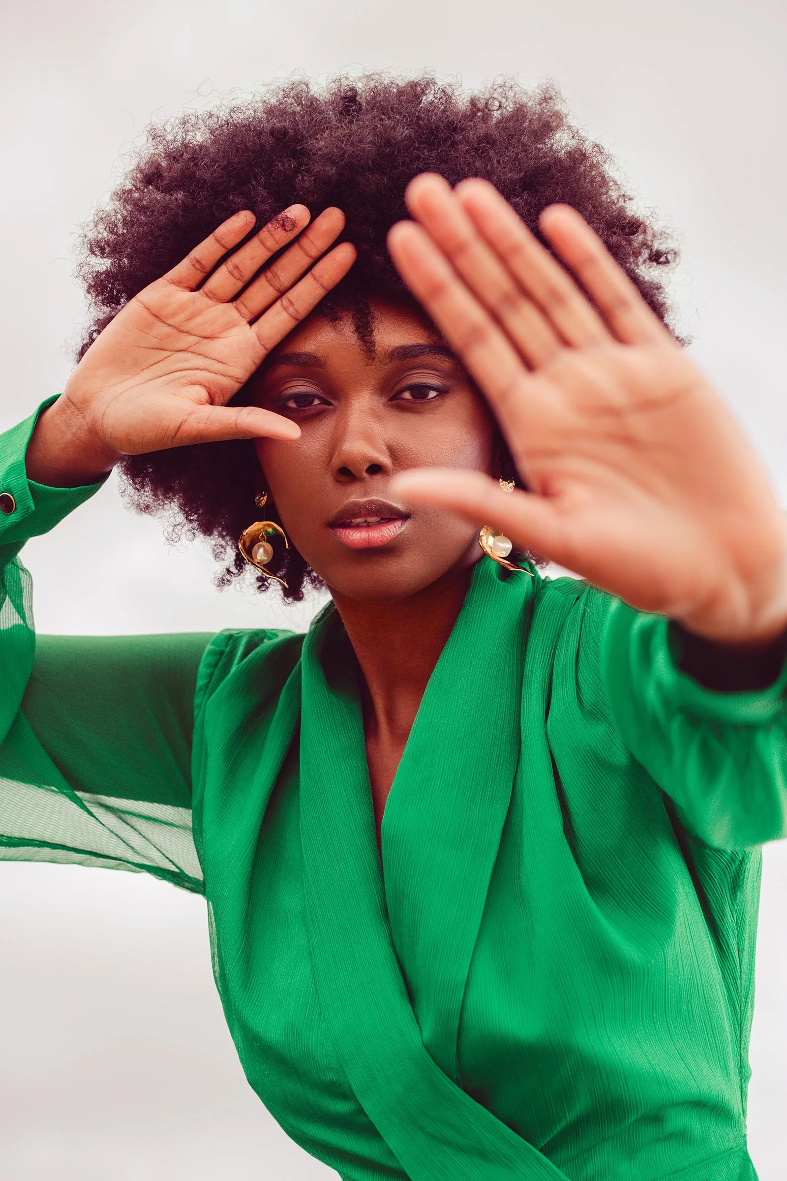 Woman in green dress posing with hands partially covering face, wearing large earrings.