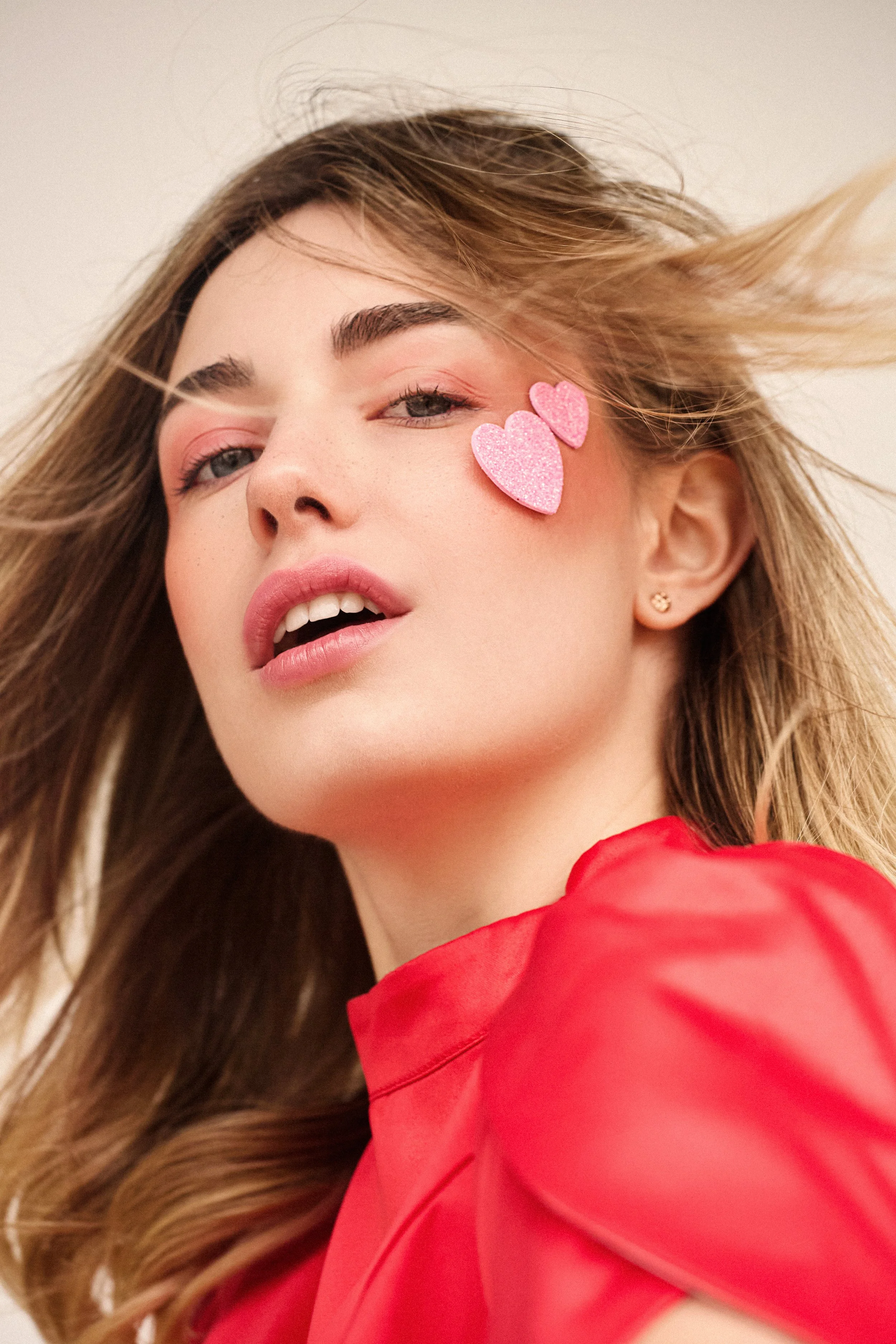 A woman with long, wavy hair and pink makeup is wearing a red outfit. She has two pink glittery heart stickers on her cheek and is looking at the camera. Her head is tilted slightly back.
