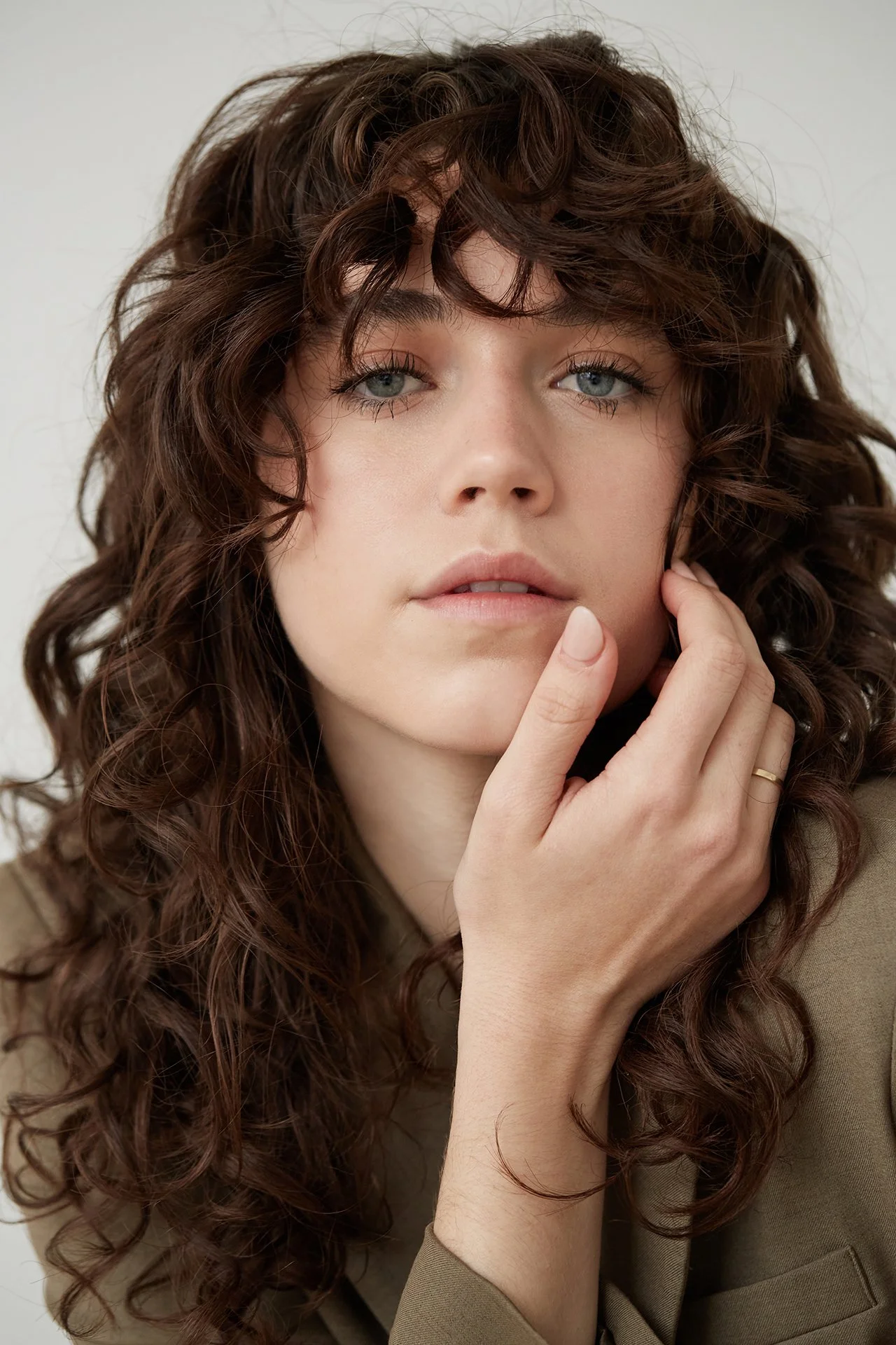 Person with curly brown hair touching their face, wearing a beige jacket.