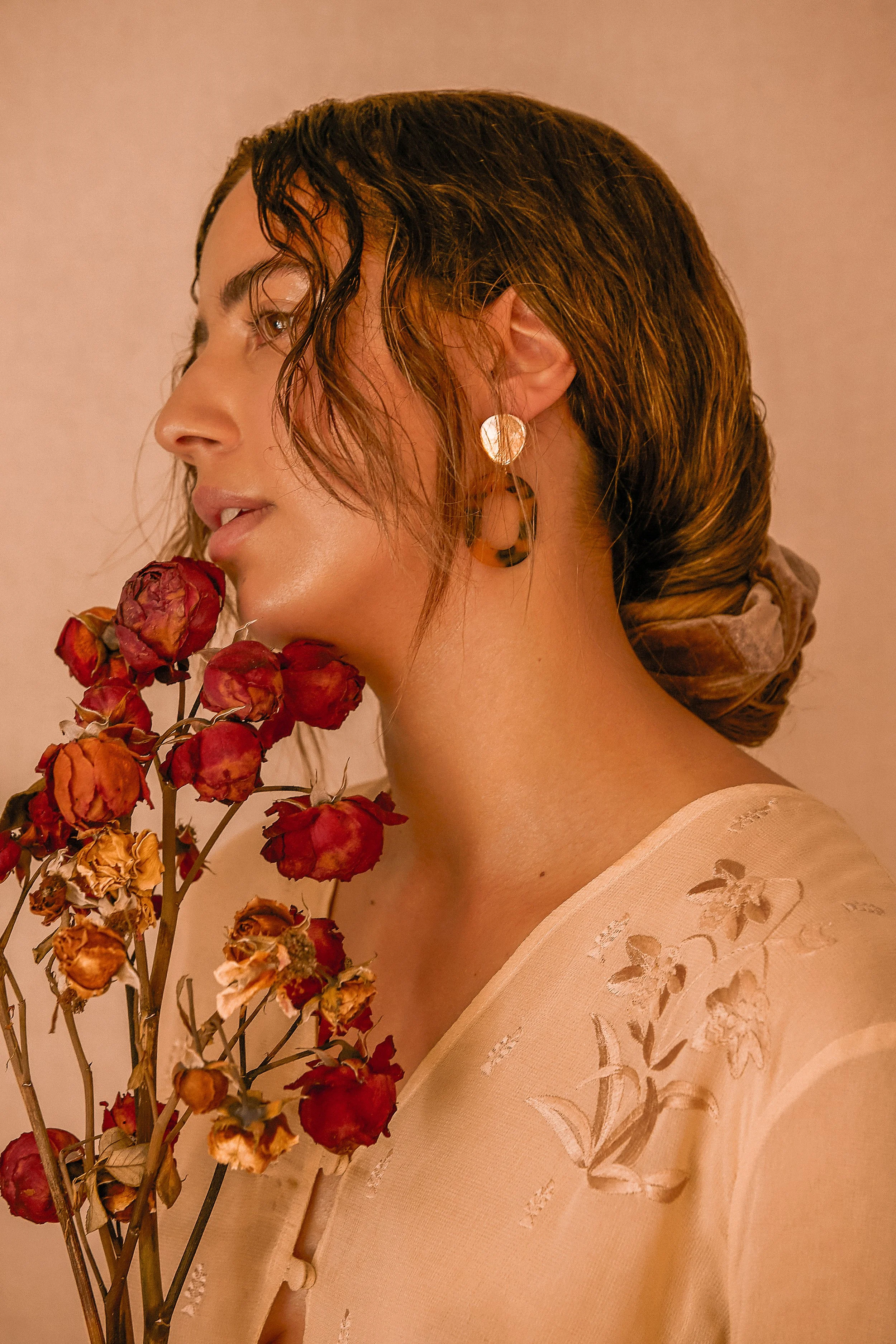 Woman with wet hair and gold earrings holding dried red roses, wearing a beige embroidered blouse.
