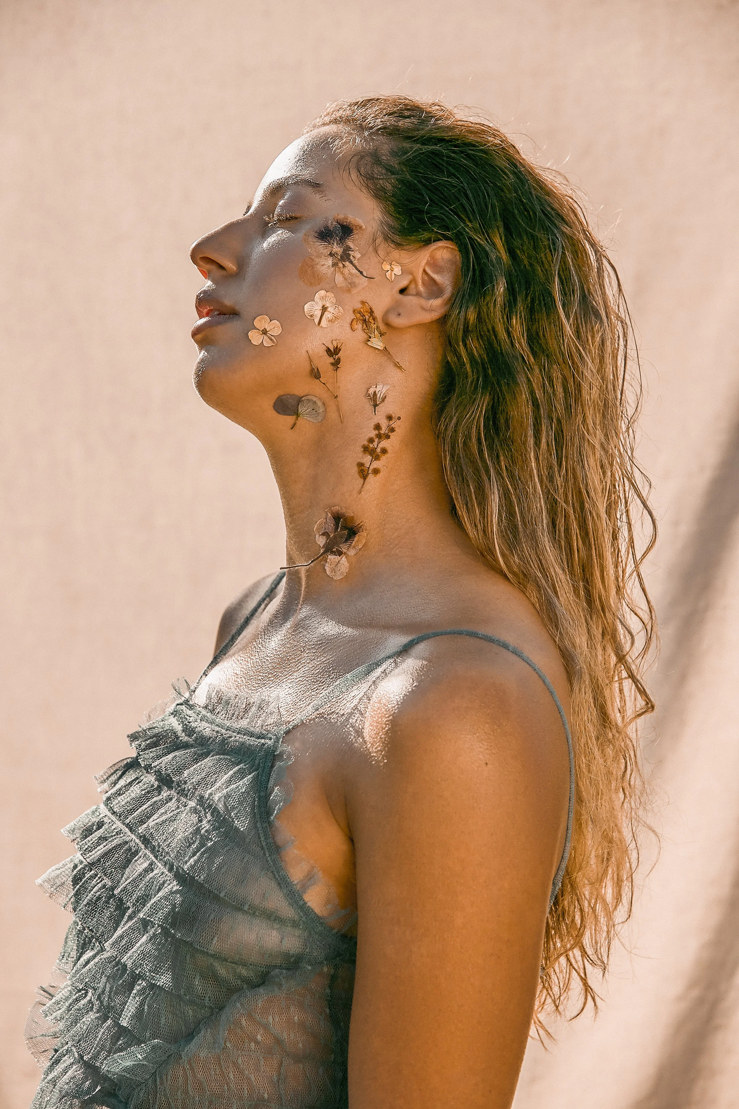 Woman with floral decorations on face, wearing sheer ruffled top, standing against neutral background.