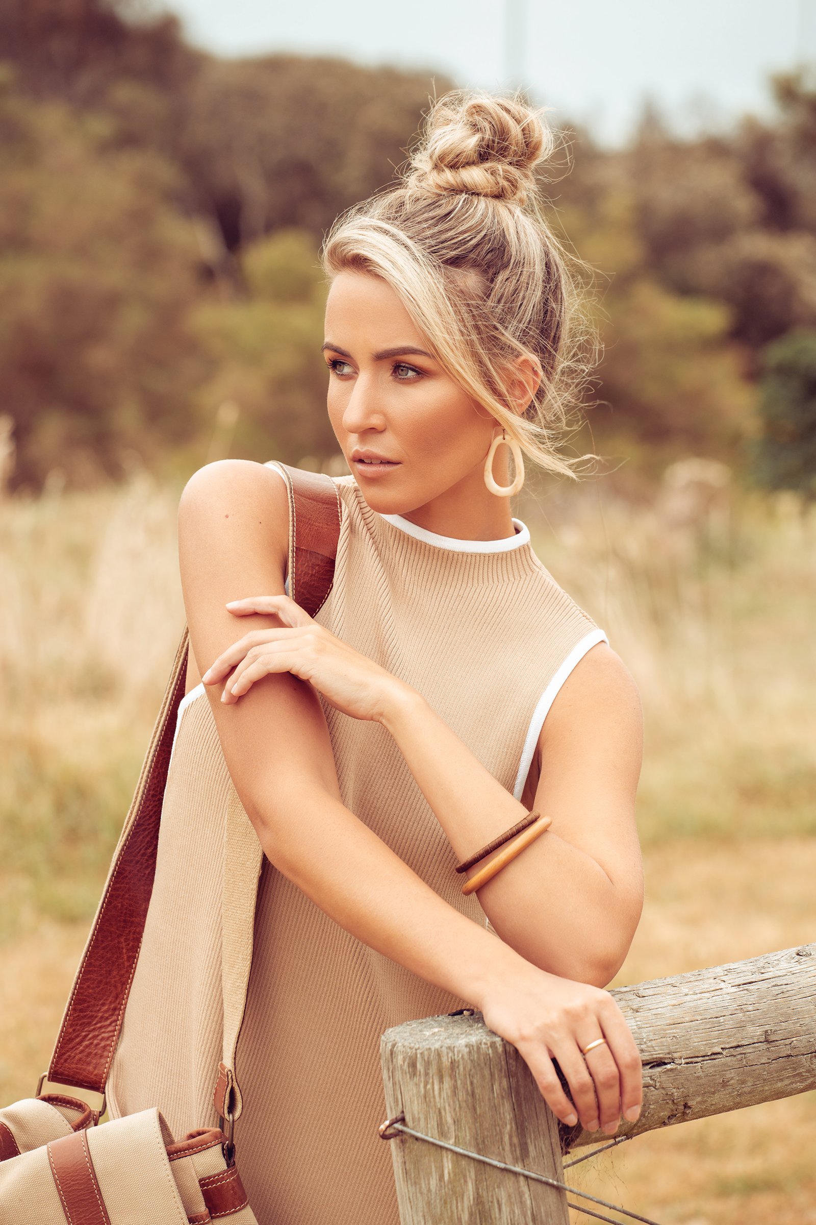 Fashionable woman with blonde hair in a topknot, wearing a beige sleeveless top, large hoop earrings, bracelets, and a bag, leaning on a wooden fence, outdoor setting.